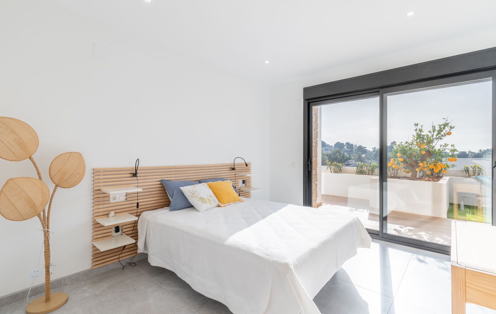 Chambre moderne avec tête de lit en lattes de bois, étagères intégrées et grandes baies vitrées ouvrant sur une terrasse avec un oranger.