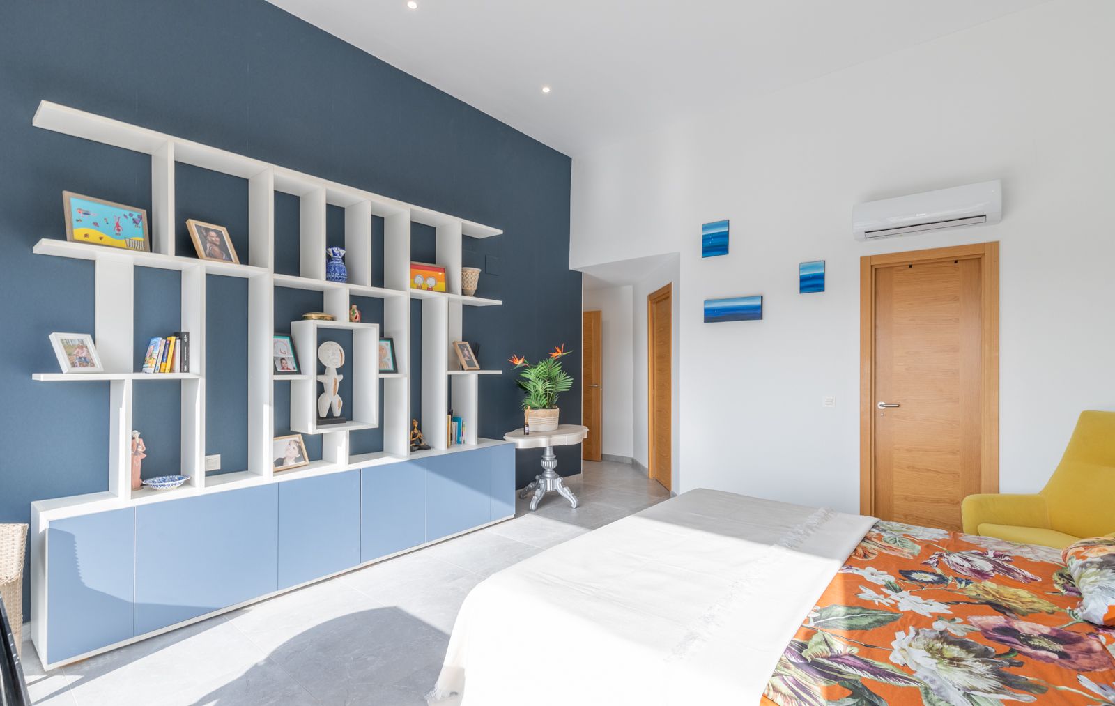 Chambre moderne avec une étagère géométrique blanche sur un mur bleu marine, des rangements intégrés et un sol en grès cérame clair.