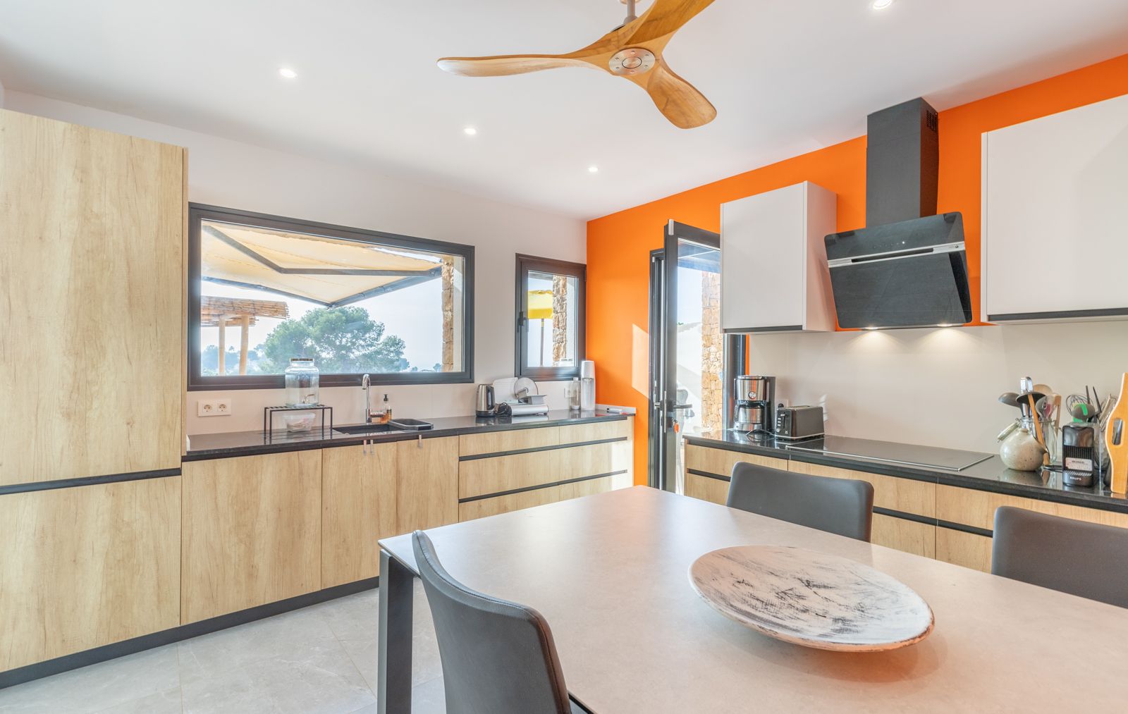 Cuisine contemporaine avec meubles en bois clair, plans de travail noirs et mur orange. Elle dispose d'un ventilateur de plafond en bois et de larges fenêtres sur l'extérieur.
