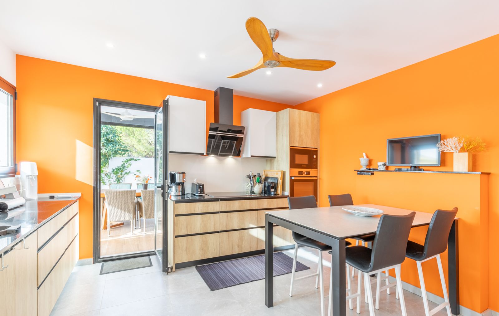 Cuisine moderne aux murs orange vif, meubles en bois clair et coin repas intégré offrant un accès direct à la terrasse extérieure.