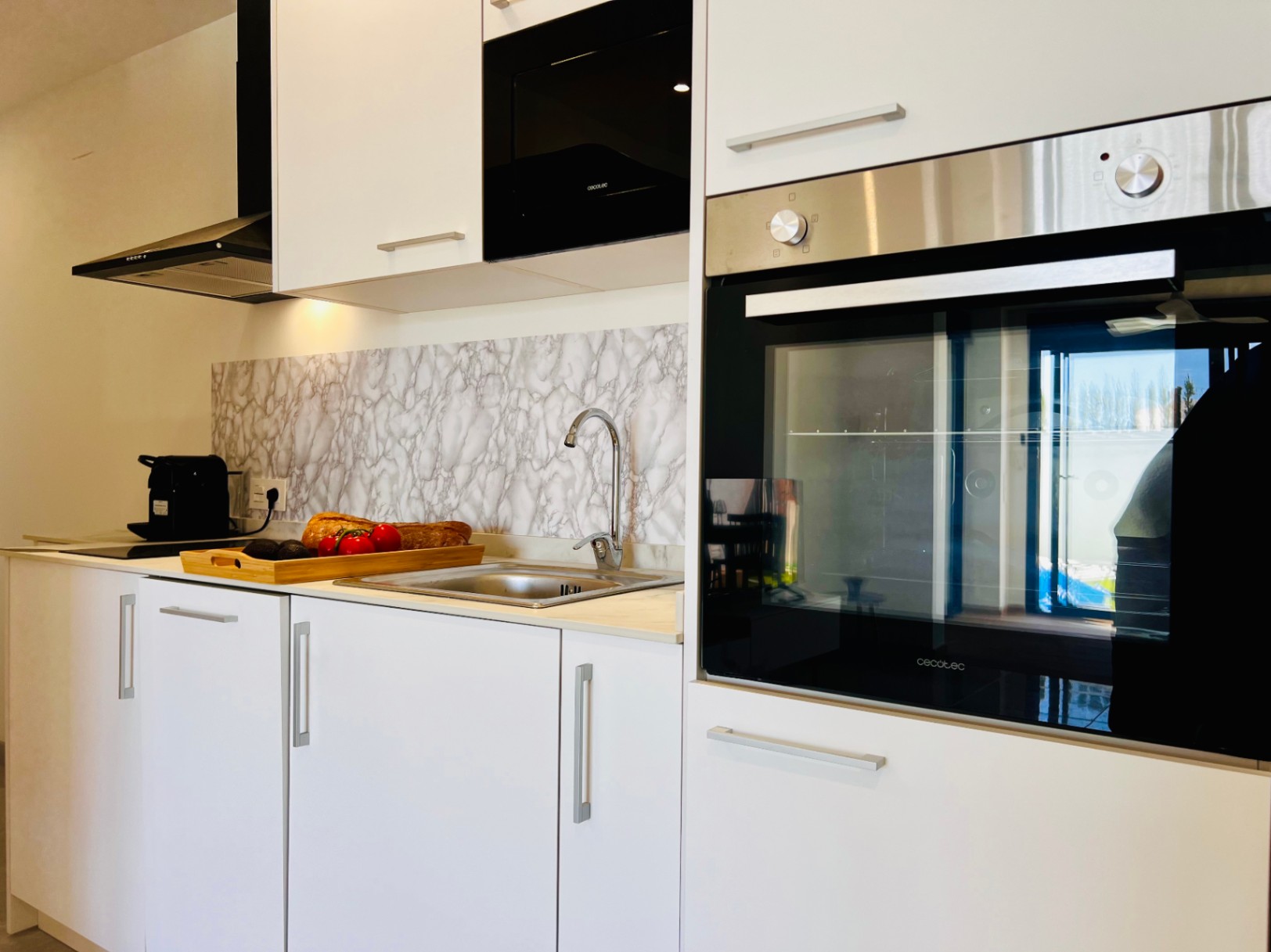 Cuisine moderne avec placards blancs, appareils Cecotec intégrés et crédence effet marbre. Elle comprend un évier en inox et une hotte aspirante noire élégante.