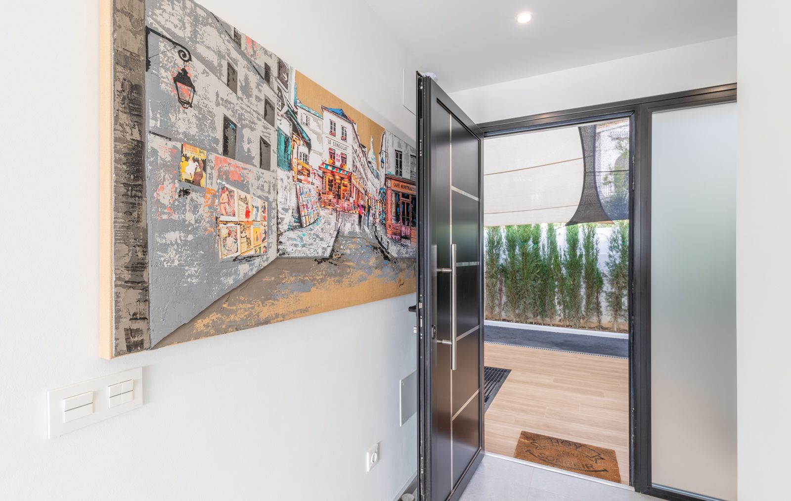 Entrée principale avec porte de sécurité moderne, tableau urbain grand format et vue sur la terrasse extérieure en bois bordée de cyprès de confidentialité.