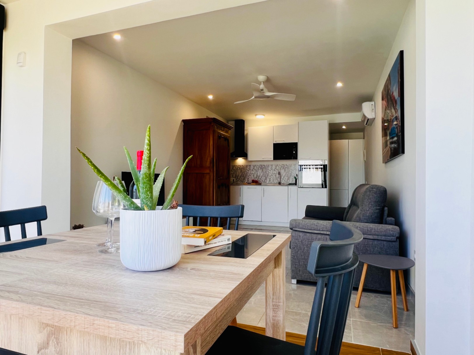 Espace de vie ouvert comprenant une cuisine blanche moderne, un canapé gris, un ventilateur de plafond et une armoire en bois classique contrastant avec le style épuré.