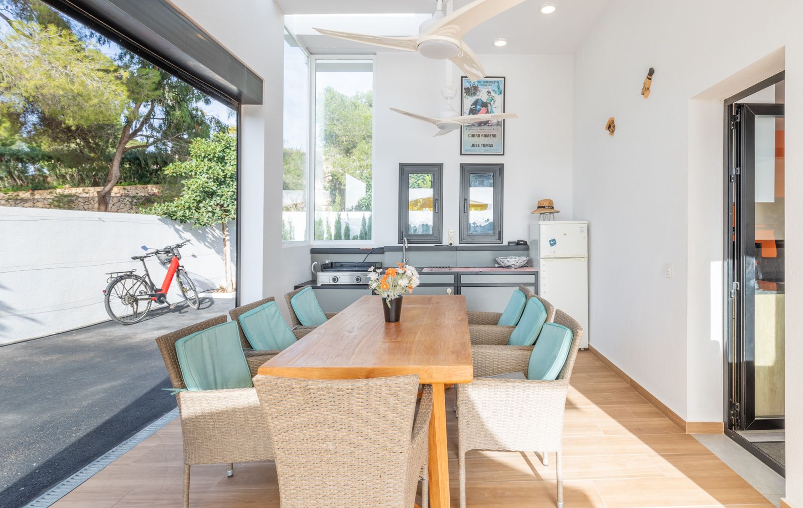 Espace repas extérieur couvert avec cuisine d'été, table en bois massif, fauteuils en osier et ventilateurs de plafond, ouvert sur la cour pavée.