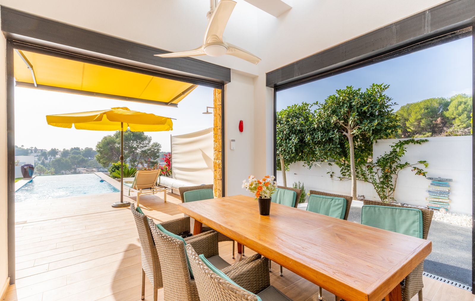 Espace repas extérieur couvert avec table en bois et chaises en osier, offrant un accès direct à la piscine à débordement et équipé d'un ventilateur de plafond.