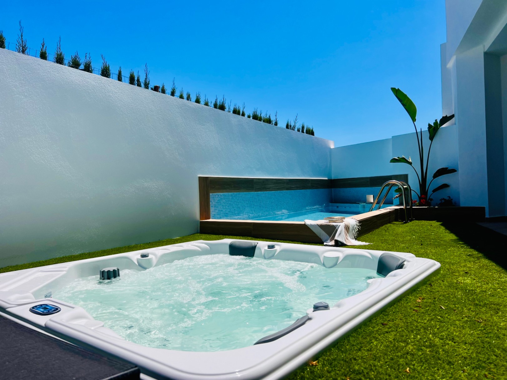 Jacuzzi extérieur encastré dans du gazon synthétique, situé à côté d'une piscine privée avec mosaïque bleue et murs d'intimité blancs.