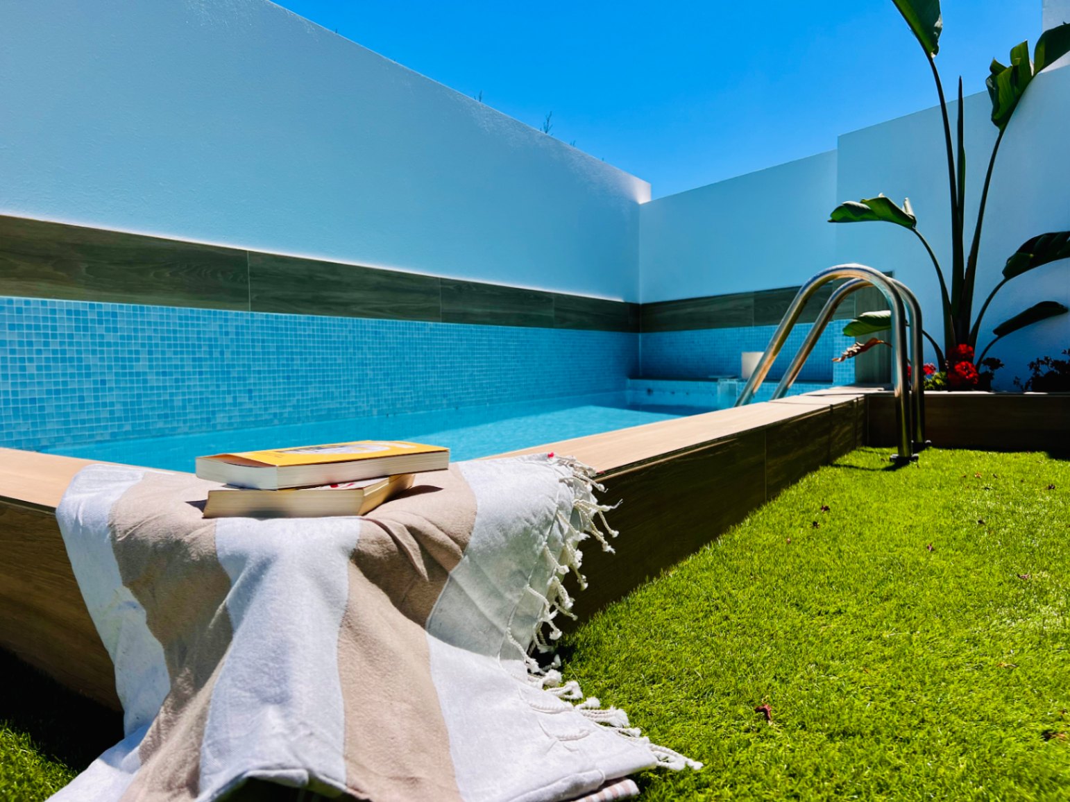Jacuzzi privé avec mosaïque bleue, bordure en bois et zone de gazon synthétique, encadré par des murs blancs épurés.