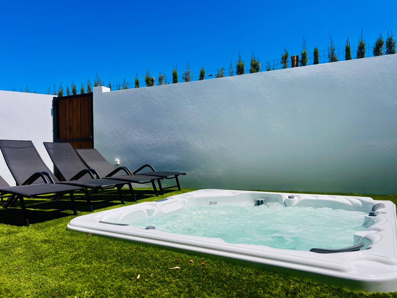 Jacuzzi privé encastré dans une pelouse synthétique, accompagné de chaises longues et d'un mur d'enceinte blanc assurant une discrétion totale.