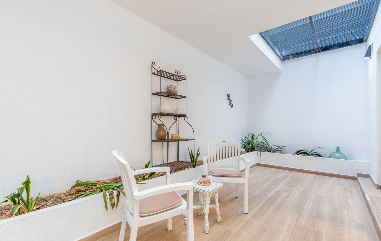 Patio intérieur lumineux avec puits de lumière, sol en grès cérame imitation bois, jardinières intégrées et coin salon avec mobilier blanc et étagère en fer forgé.