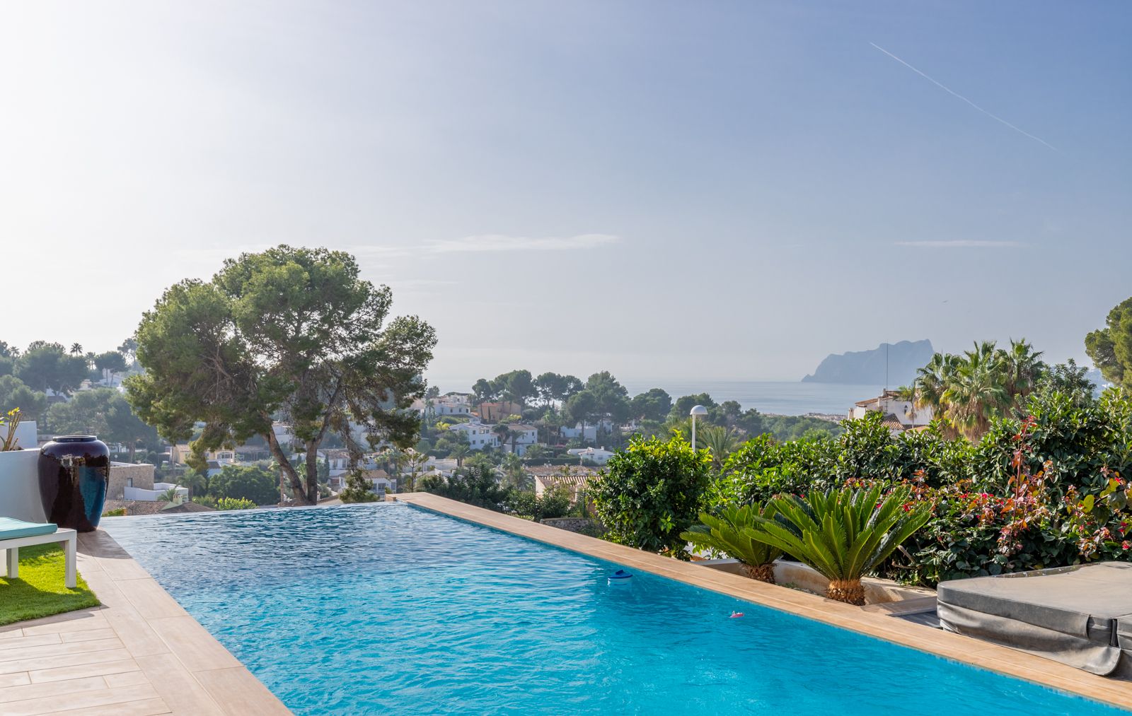 Piscine à débordement avec vue panoramique sur la mer Méditerranée et le Peñón de Ifach, entourée d'une terrasse en pierre claire et d'un jardin arboré.