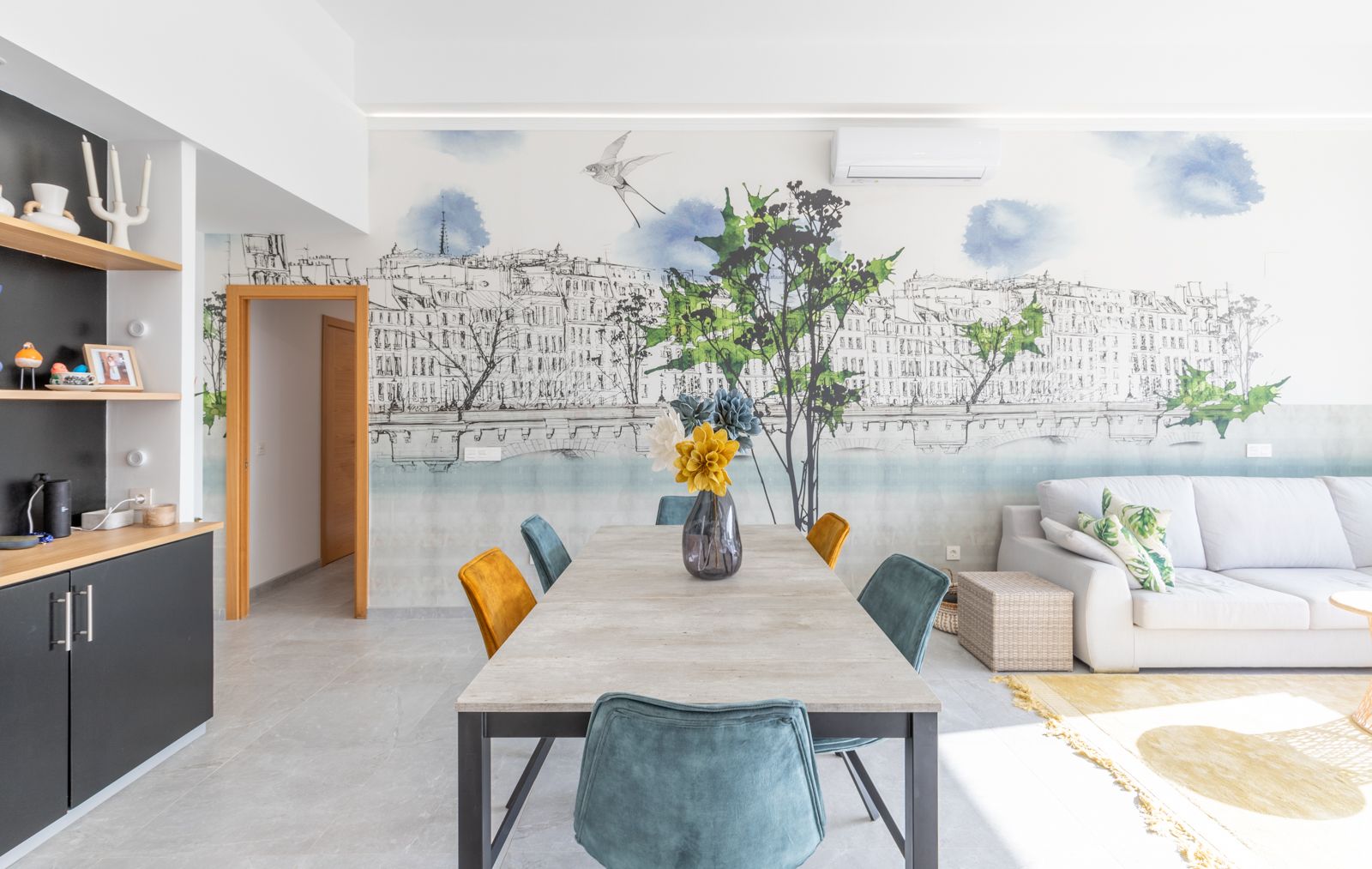 Salle à manger moderne avec table effet pierre, chaises en velours bleu et moutarde, et une fresque murale urbaine. Sols en grès gris et salon adjacent.