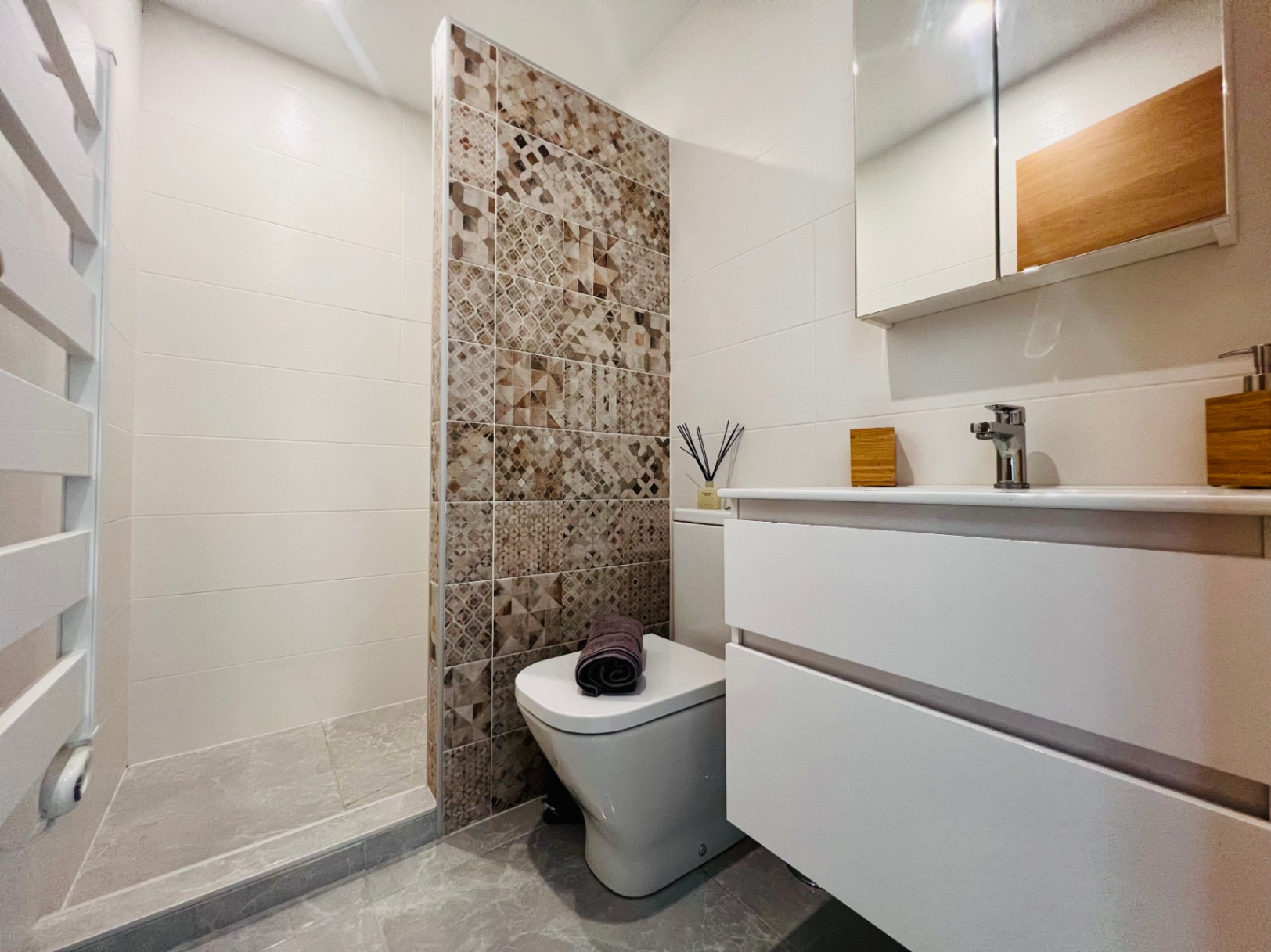 Salle de bain moderne avec douche à l'italienne, mur décoratif en carreaux géométriques, meuble vasque blanc et radiateur sèche-serviettes.