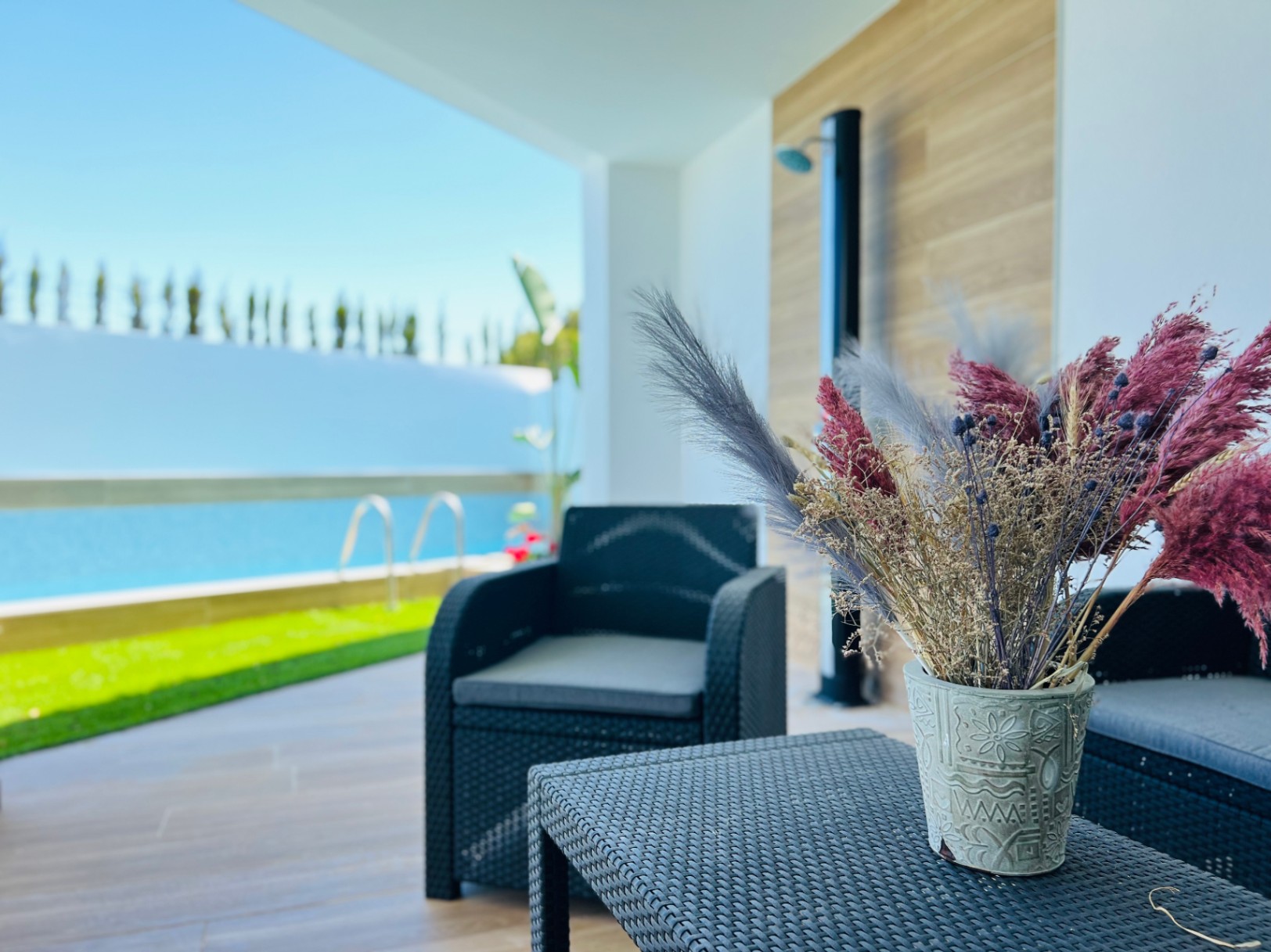 Salon de terrasse ombragé avec mobilier en rotin synthétique noir, offrant une vue dégagée sur la piscine et les espaces verts extérieurs.