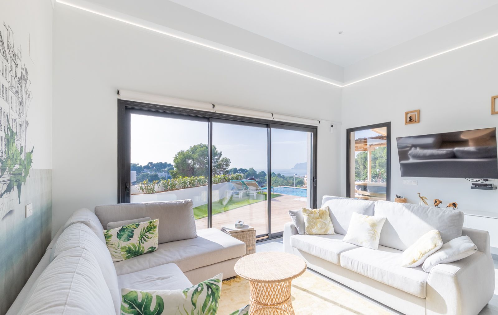 Salon lumineux avec canapés gris, éclairage LED intégré et grandes baies vitrées ouvrant sur la terrasse, la piscine et une vue dégagée sur la mer.
