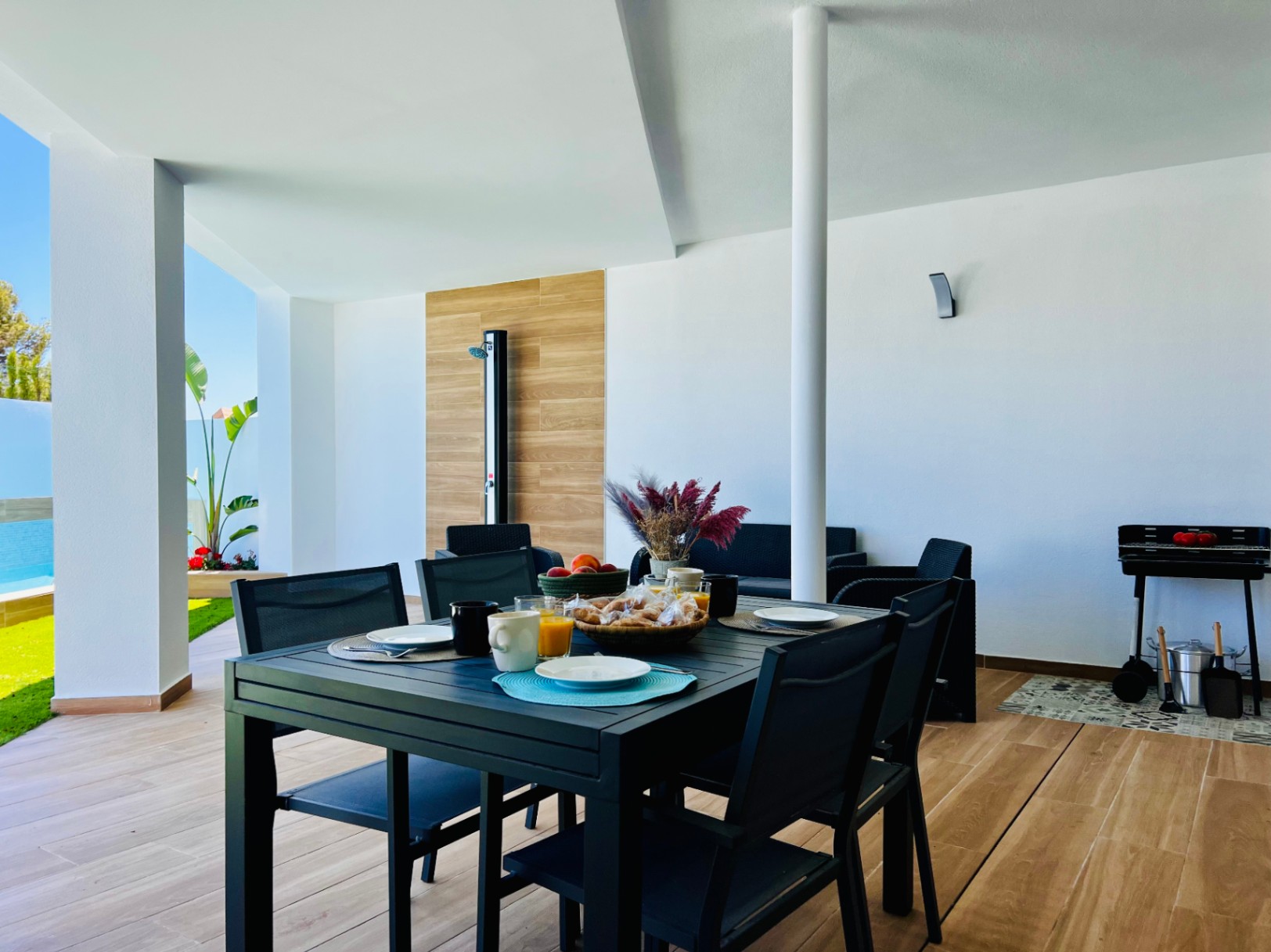 Terrasse couverte avec sol en grès cérame aspect bois, espace repas extérieur, barbecue et douche extérieure intégrée sur panneau décoratif.
