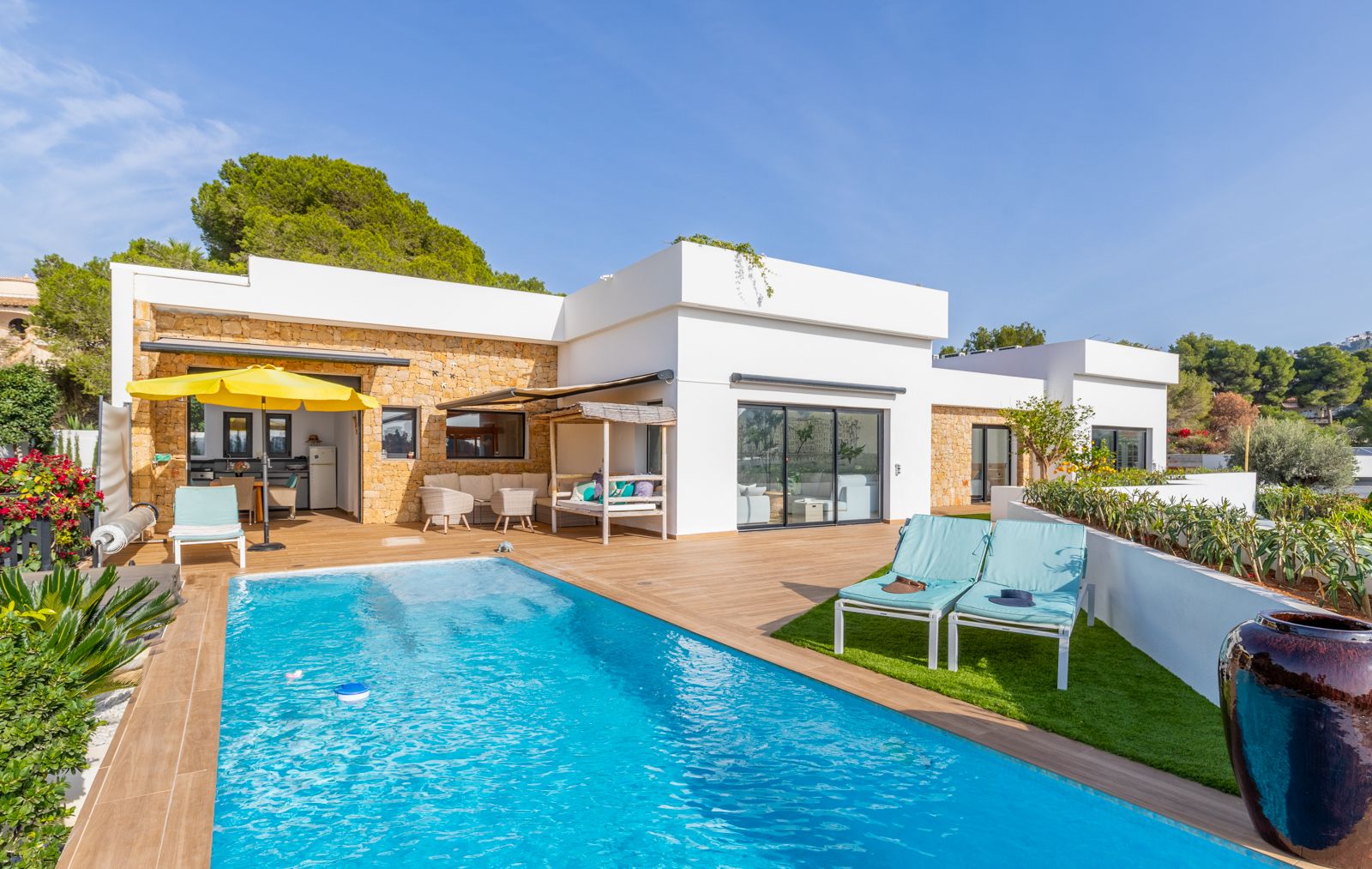 Terrasse extérieure avec piscine, sol en grès cérame imitation bois et murs en pierre naturelle. Comprend une cuisine d'été, un solarium en gazon synthétique et des coins salon.