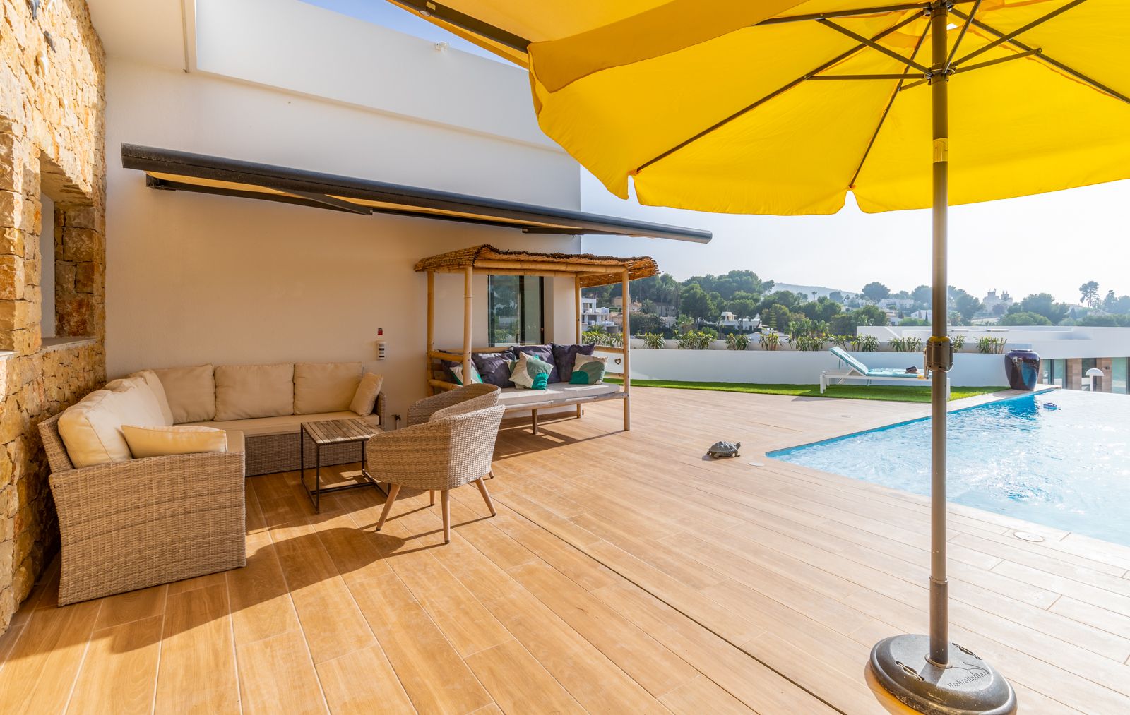 Terrasse extérieure avec sol en grès cérame imitation bois, salon de jardin en osier, lit balinais en bambou et piscine bordée d'un mur en pierre naturelle.