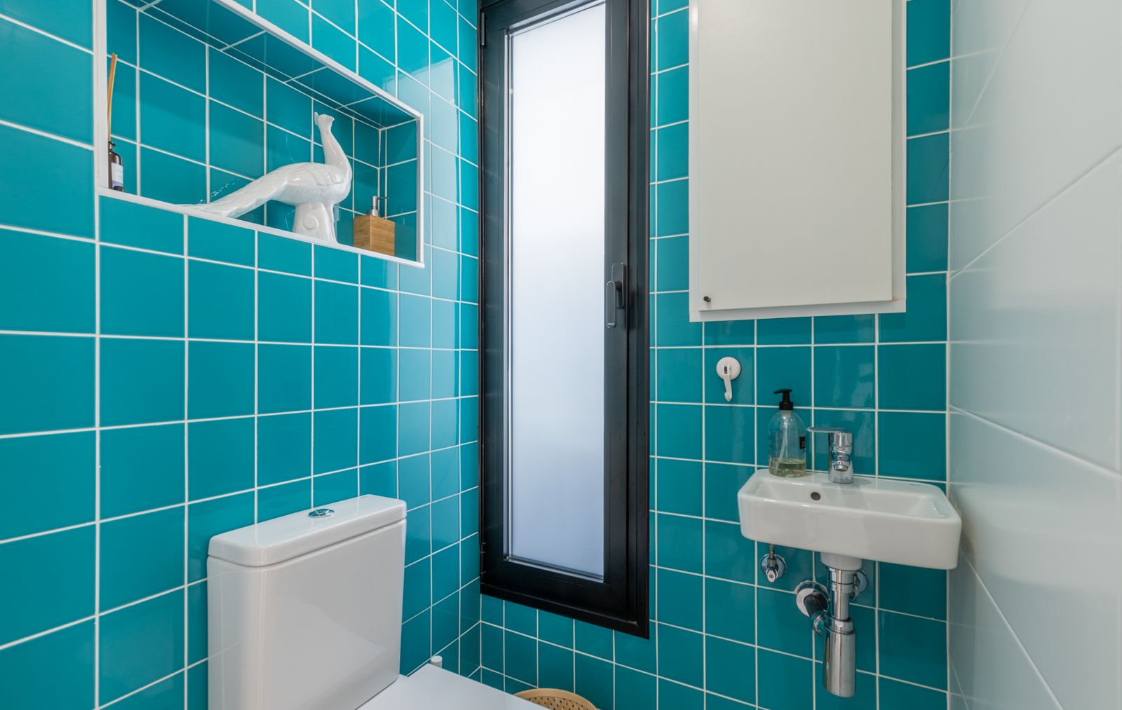 Toilettes invités avec carrelage turquoise éclatant, niche murale intégrée et petit lavabo suspendu. Une fenêtre étroite en verre dépoli assure l'intimité.