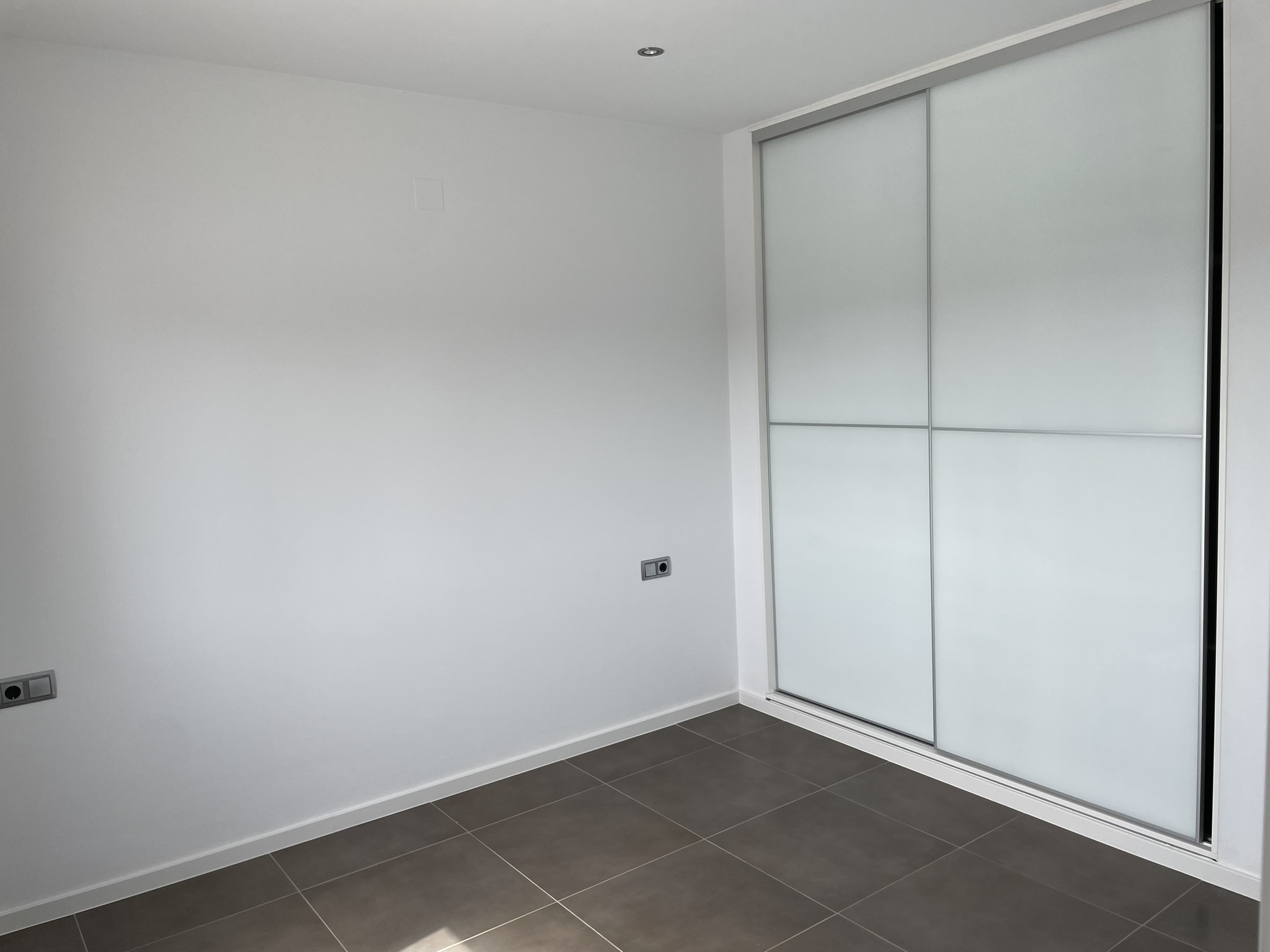 Chambre avec placard intégré à portes coulissantes blanches, carrelage au sol de ton sombre et éclairage par spots encastrés.