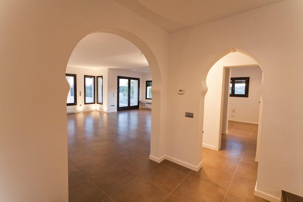 Couloir avec arches de style méditerranéen, sol en grès cérame teinté terre et vue sur le salon doté de menuiseries extérieures sombres.