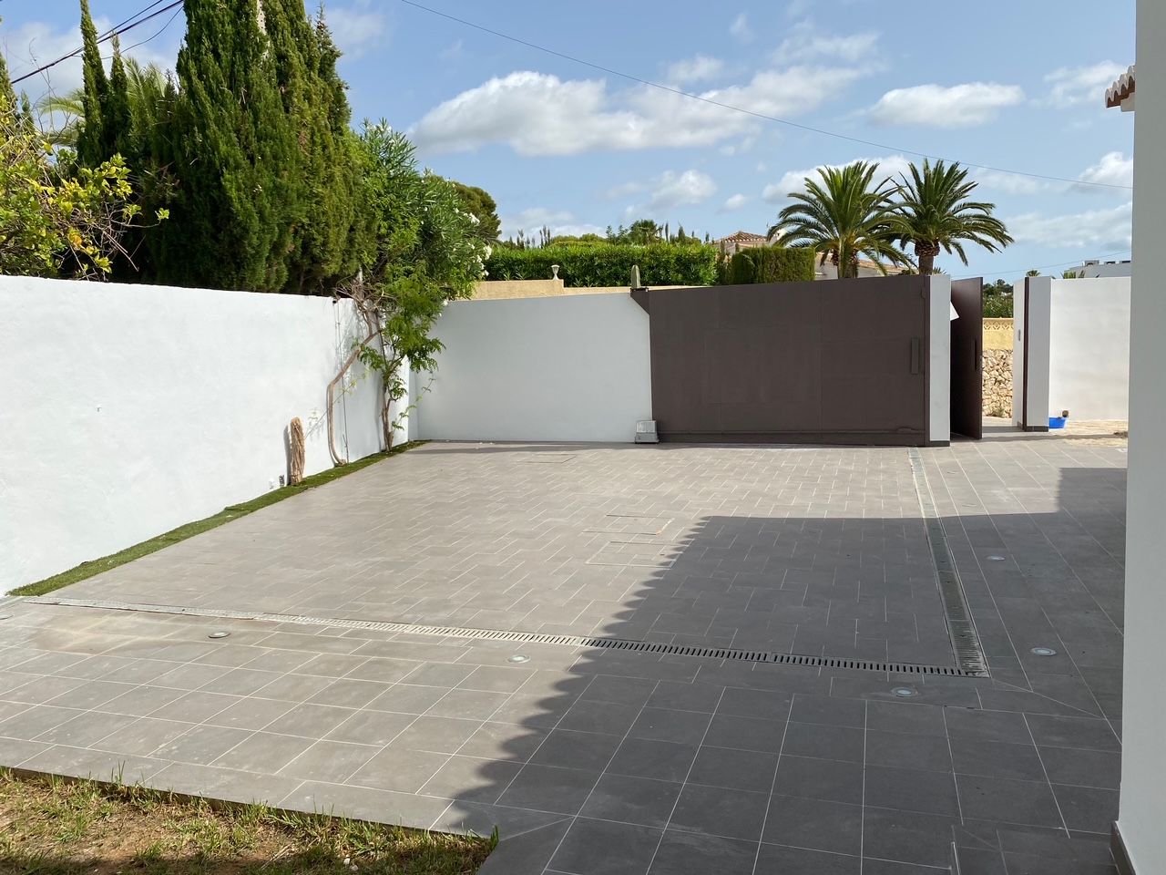 Espace de stationnement privé avec revêtement en dalles grises, portail coulissant motorisé et accès piéton, encadré par des murs blancs et des cyprès.