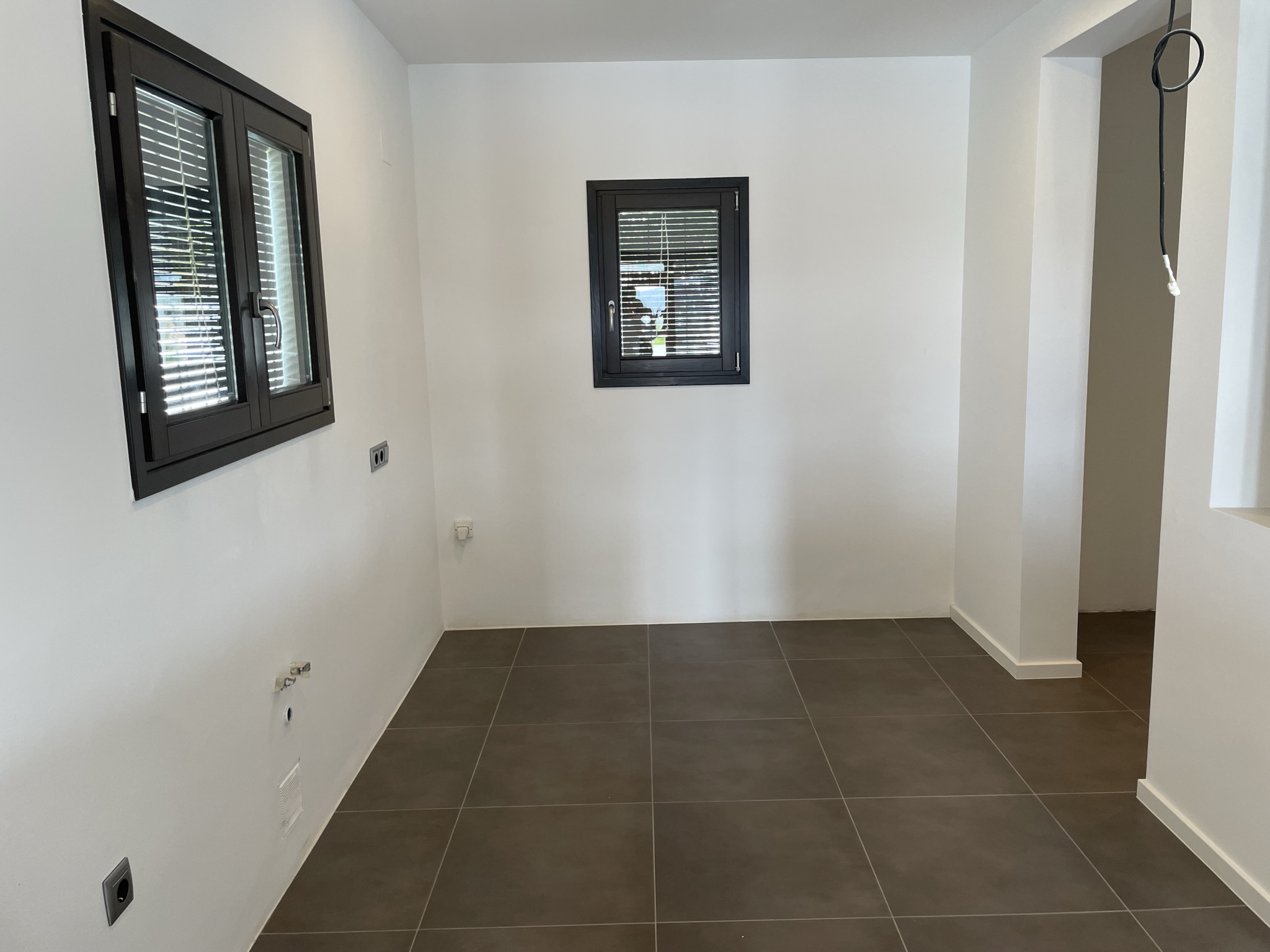Espace intérieur avec pré-installations de plomberie et d'électricité, sol en grès cérame foncé et fenêtres en aluminium avec stores intégrés.