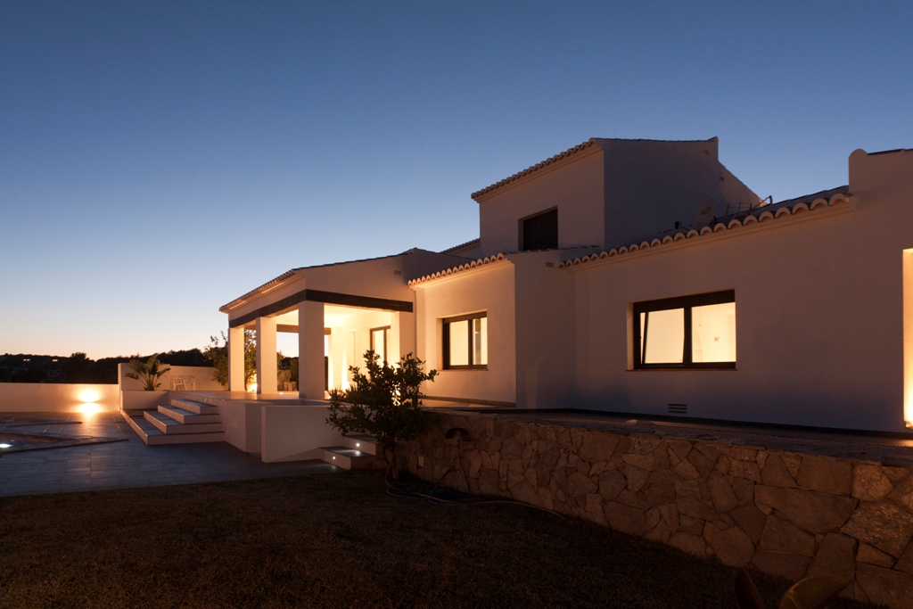 Façade extérieure de la villa au crépuscule, mettant en valeur l'éclairage chaleureux, les murs en pierre naturelle et les terrasses menant à la piscine.