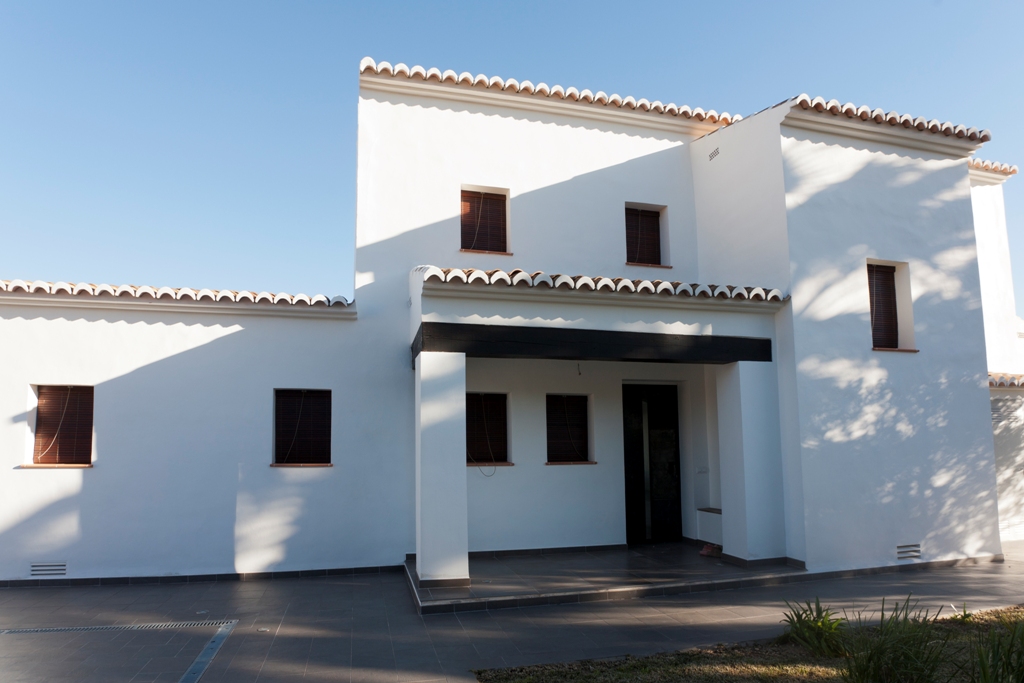 Façade méditerranéenne aux murs blancs, menuiseries en aluminium imitation bois et toiture en tuiles traditionnelles avec un porche d'entrée ombragé.