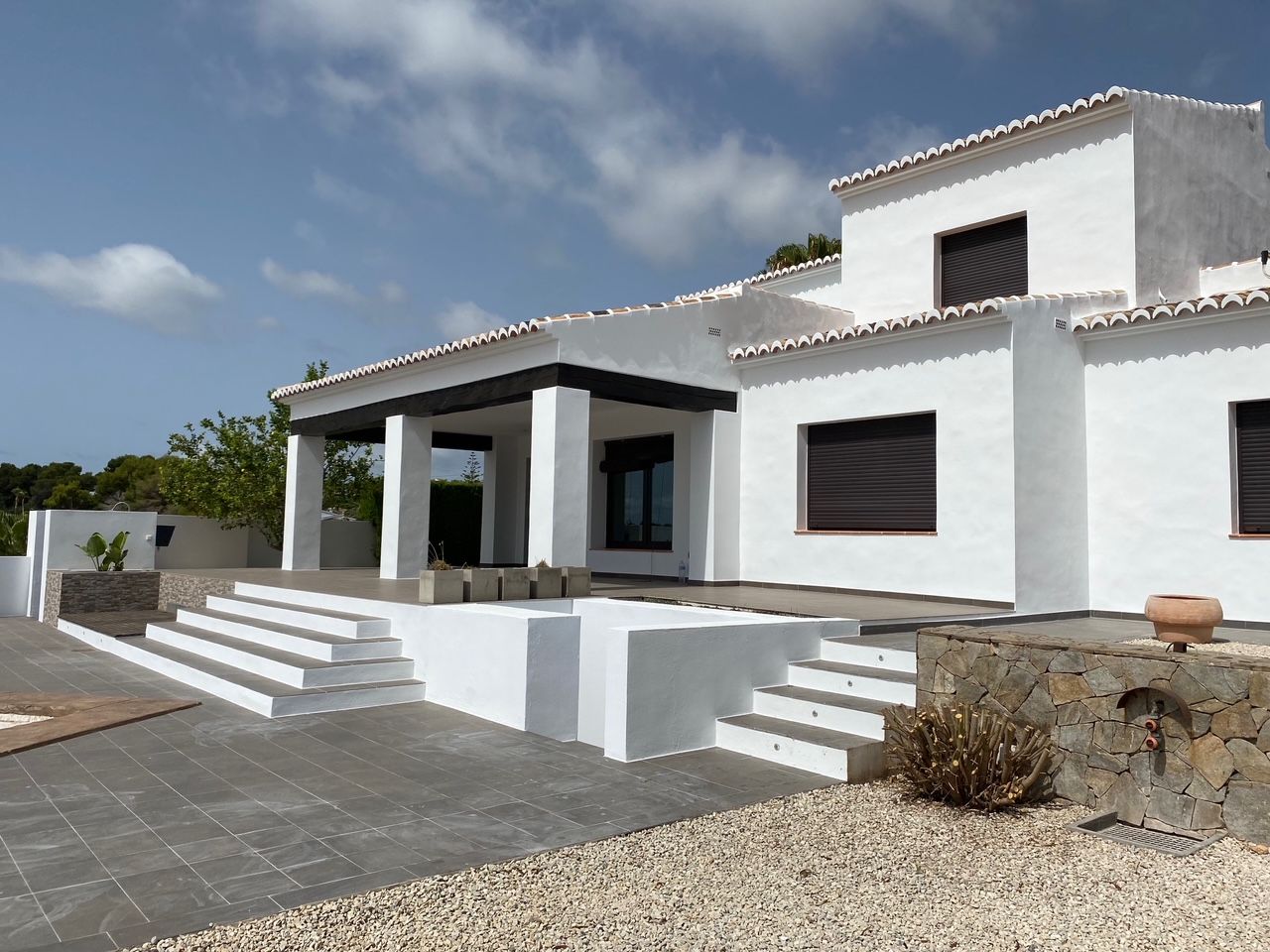 Façade méditerranéenne blanche avec poutres en bois sombre, grande terrasse carrelée et murs en pierre naturelle. Comprend un espace douche extérieure et des marches d'accès.