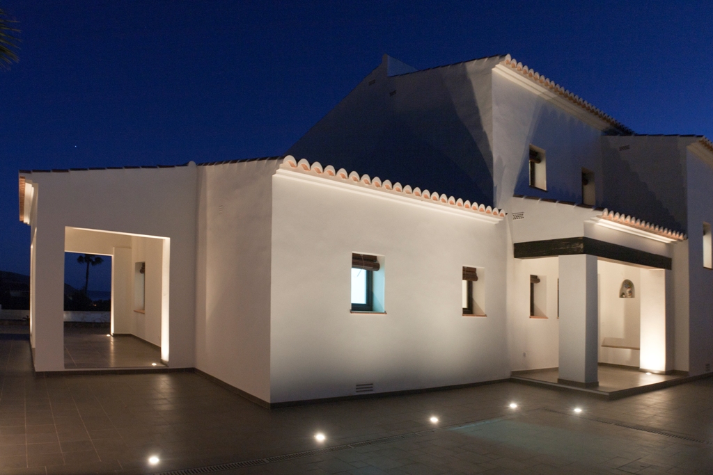 Façade méditerranéenne blanche illuminée au crépuscule par des spots encastrés au sol. Toiture en tuiles romanes et porche avec poutres sombres sur une terrasse carrelée.