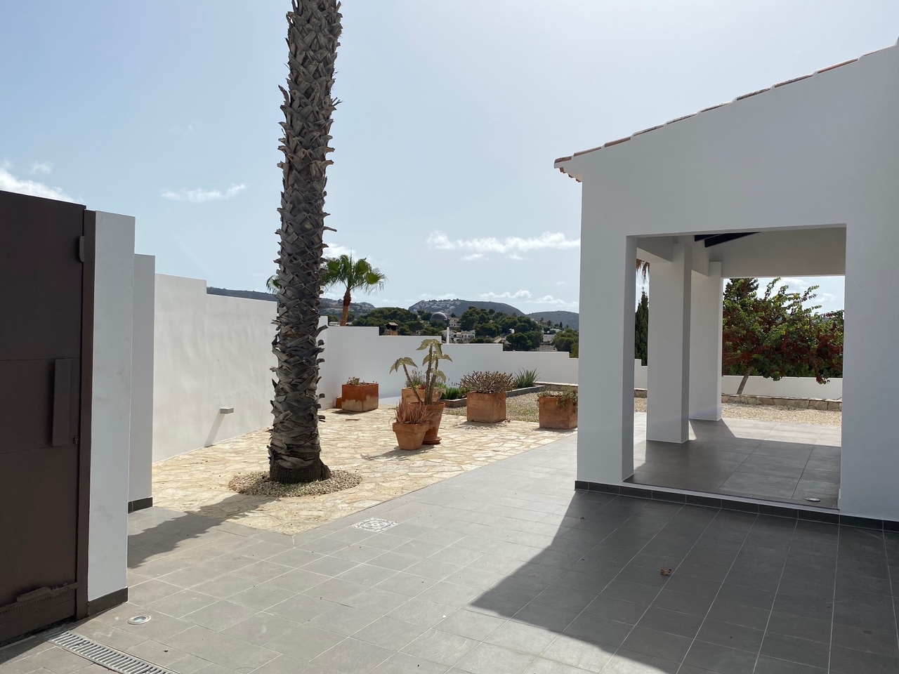 Patio extérieur avec carrelage gris et pavage en pierre naturelle, agrémenté d'un palmier et de pots en terre cuite offrant une vue sur les collines environnantes.