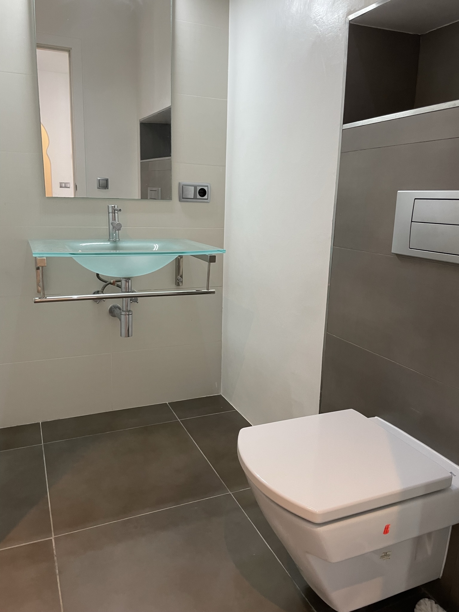 Salle de bains moderne avec vasque en verre translucide et WC suspendu. Les grands carreaux gris et crème soulignent un design épuré et fonctionnel.