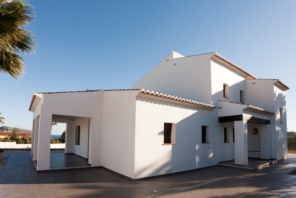 Vue extérieure de la villa méditerranéenne avec façades blanches, toits en tuiles et vastes terrasses pavées menant à un porche couvert avec vue sur mer.