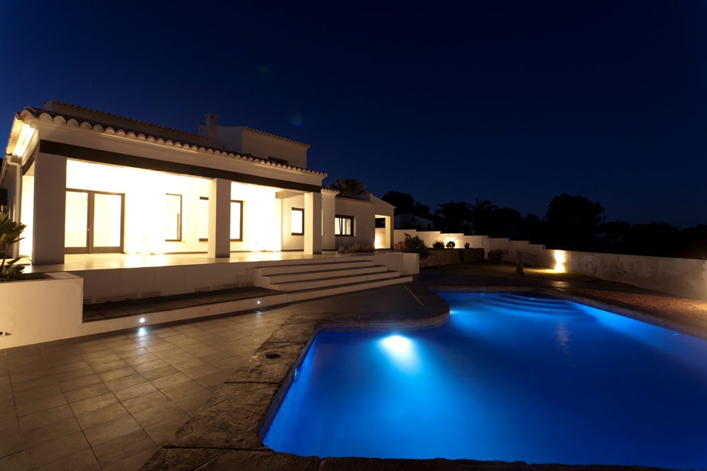 Vue nocturne de la façade blanche éclairée et de la piscine de forme irrégulière avec éclairage bleu, bordée d'une grande terrasse en grès.