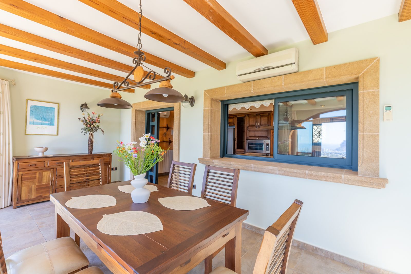 Salle à manger lumineuse avec poutres en bois et vue sur la cuisine équipée.