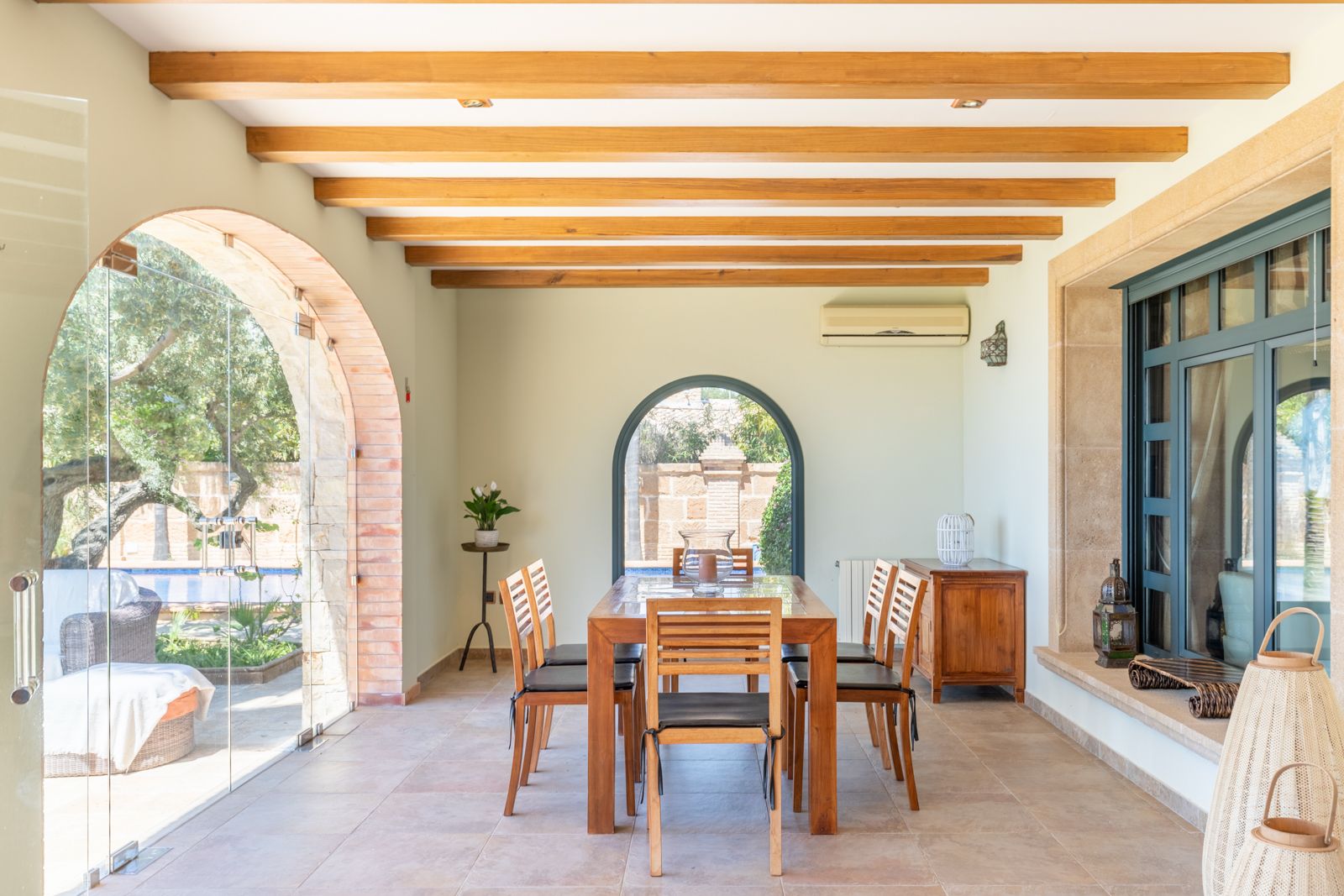 Salle à manger lumineuse avec table en bois et accès au jardin et à la piscine par des arches en brique.