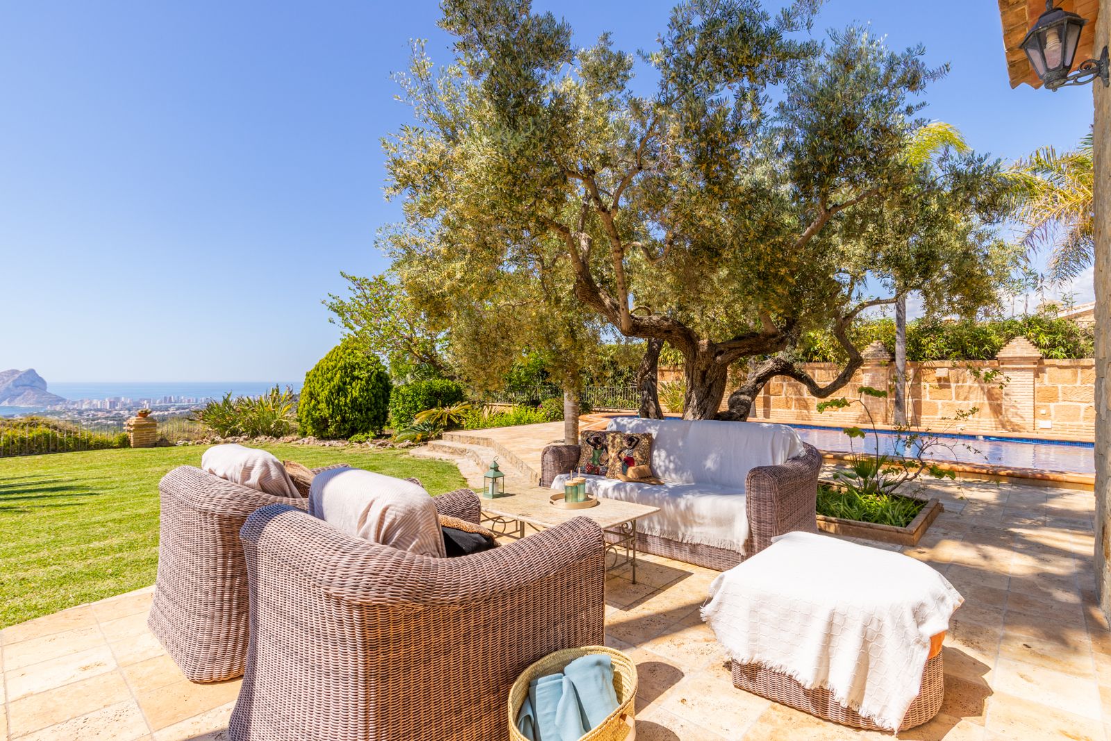 Terrasse avec mobilier en osier et vue sur mer, ombragée par un olivier centenaire.