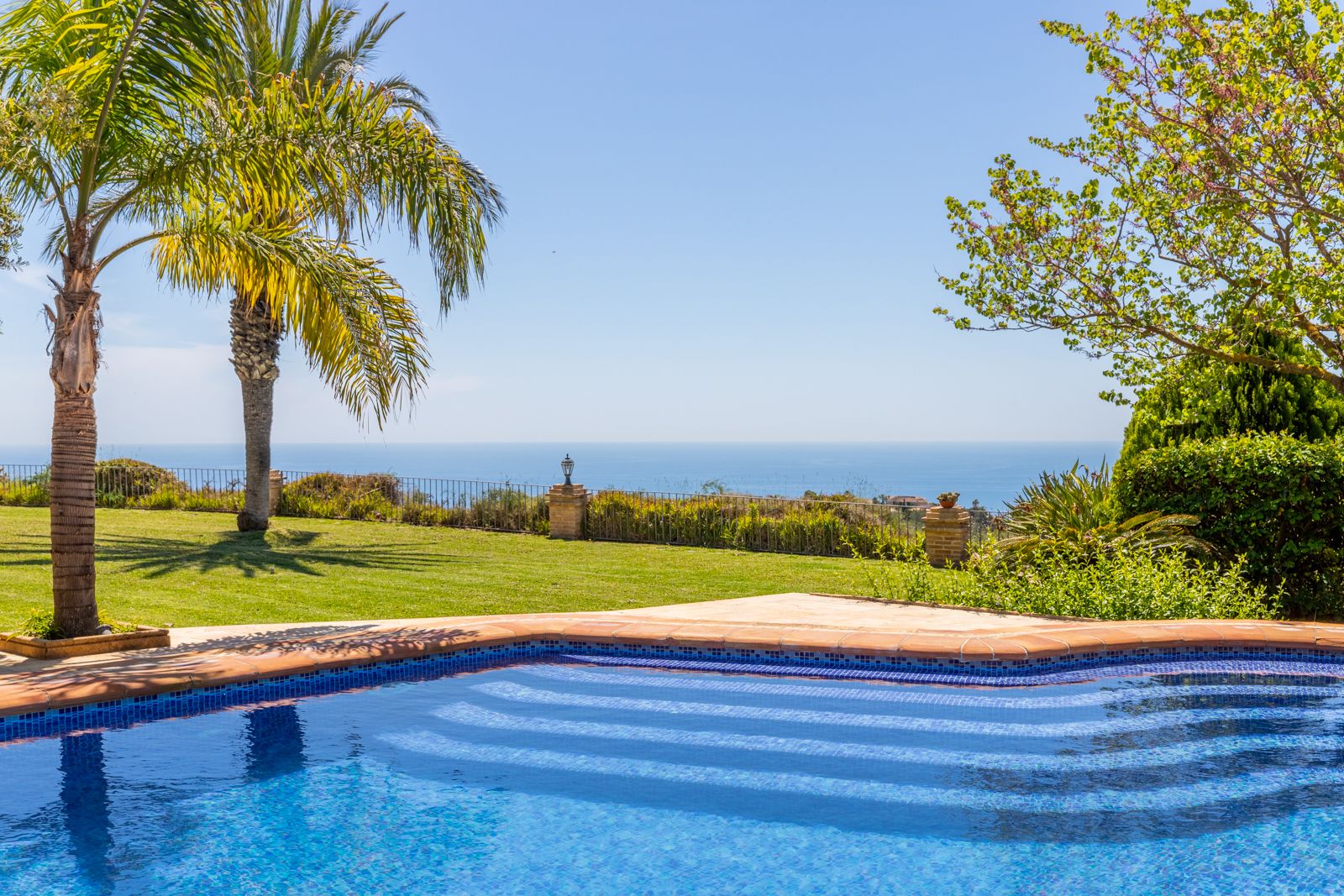 Vue sur la piscine en mosaïque bleue et le jardin avec palmiers, face à la mer Méditerranée.