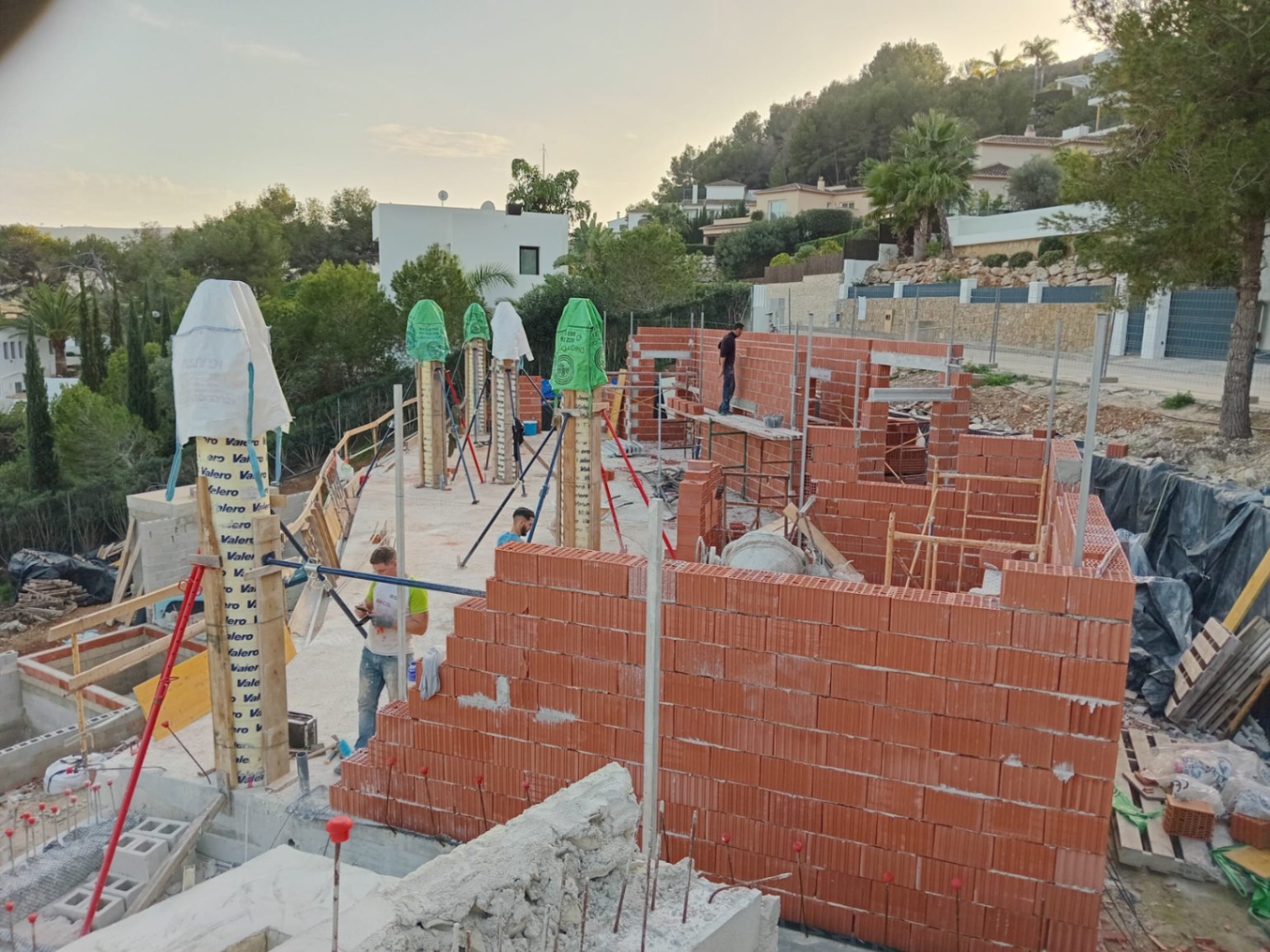 État actuel du chantier montrant l'élévation des murs en briques et des piliers structurels sur un terrain en pente avec vue sur les collines environnantes.