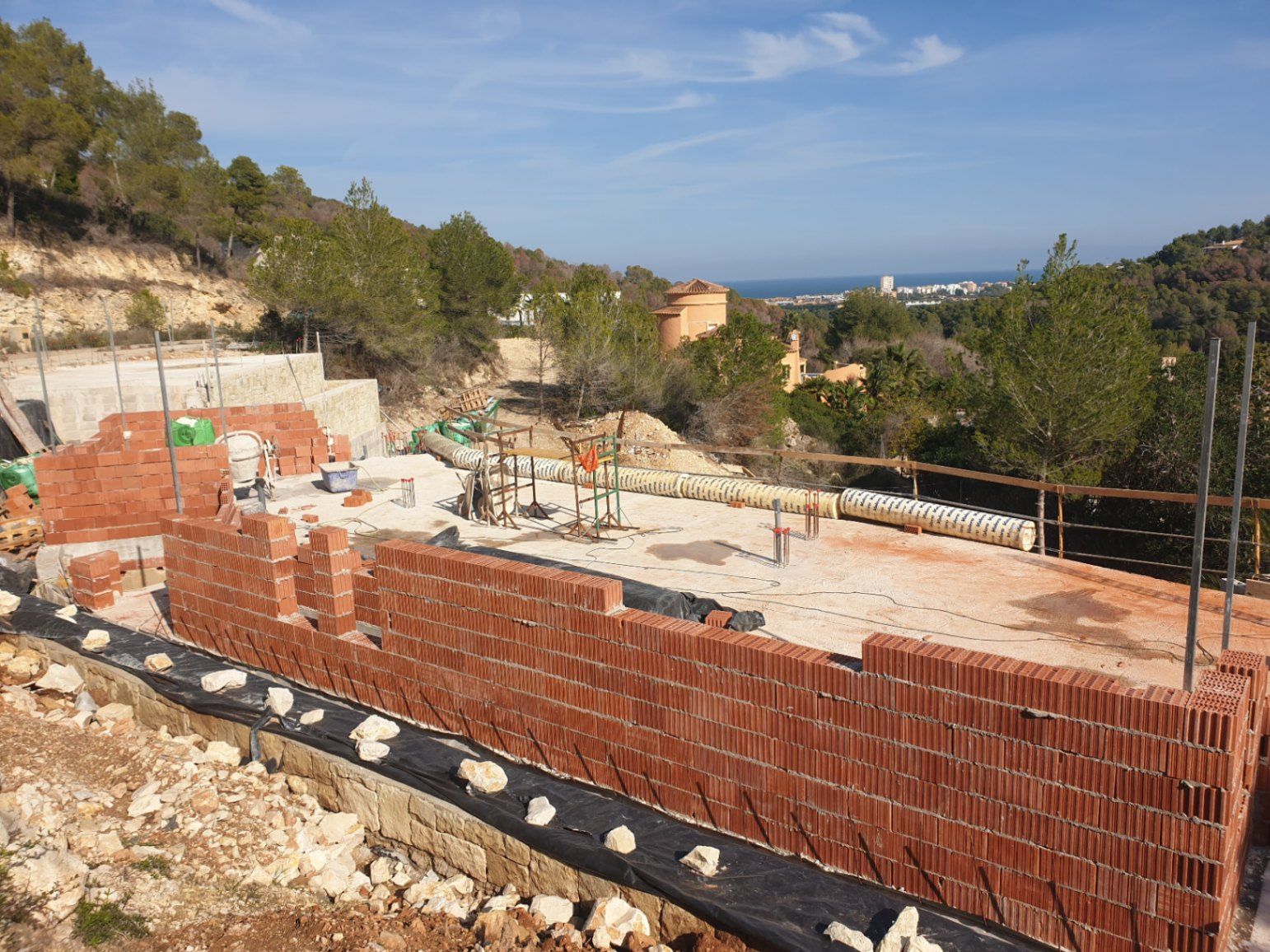 Phase de construction actuelle montrant l'élévation des murs en briques sur la dalle, avec une vue dégagée sur la mer Méditerranée et les collines environnantes.