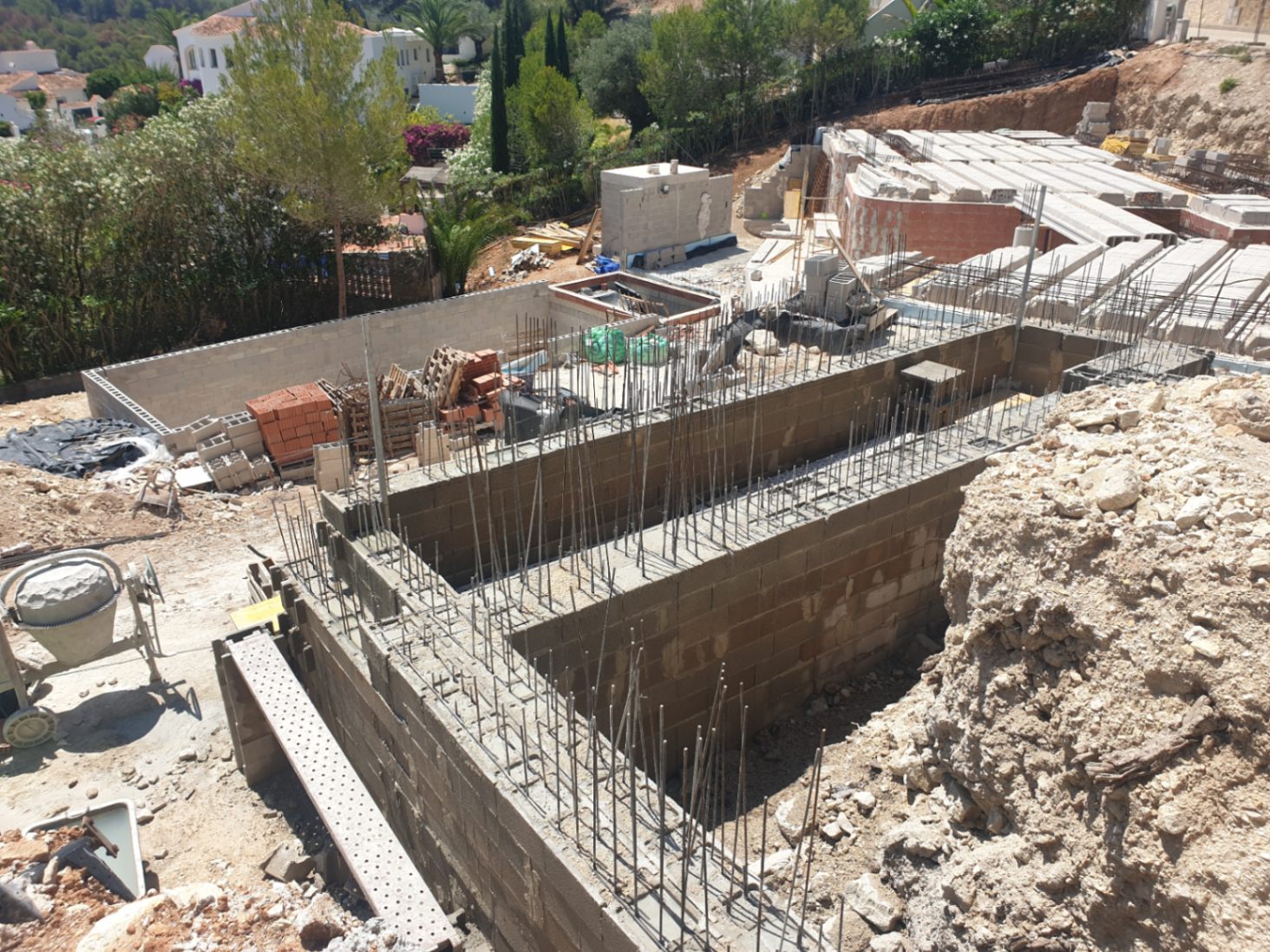 Phase initiale de construction montrant les murs de fondation avec armatures en acier et blocs de béton sur un terrain surélevé en pleine nature.