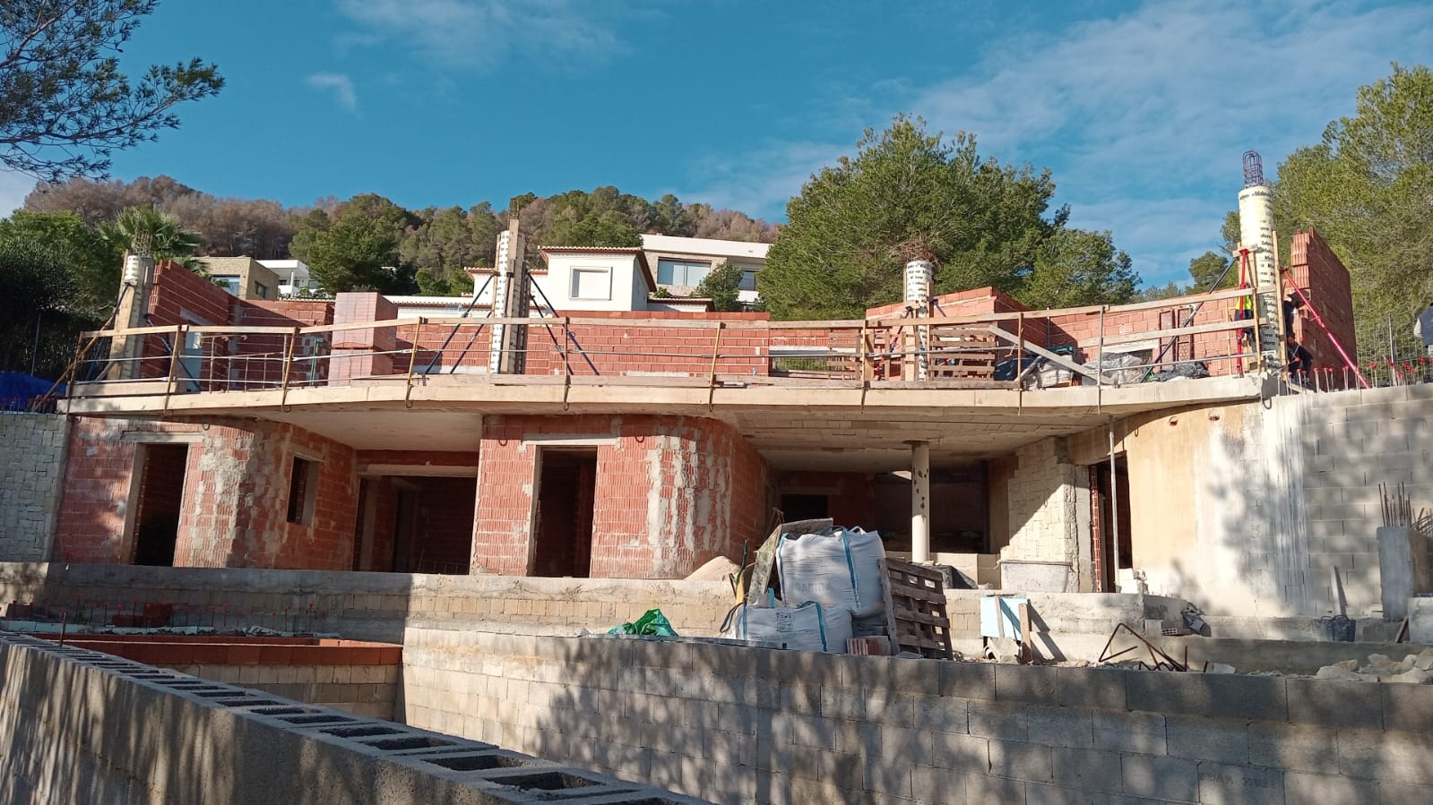 Vue de la structure en cours de construction, illustrant la terrasse supérieure courbe et les niveaux en briques rouges sur un terrain en pente.