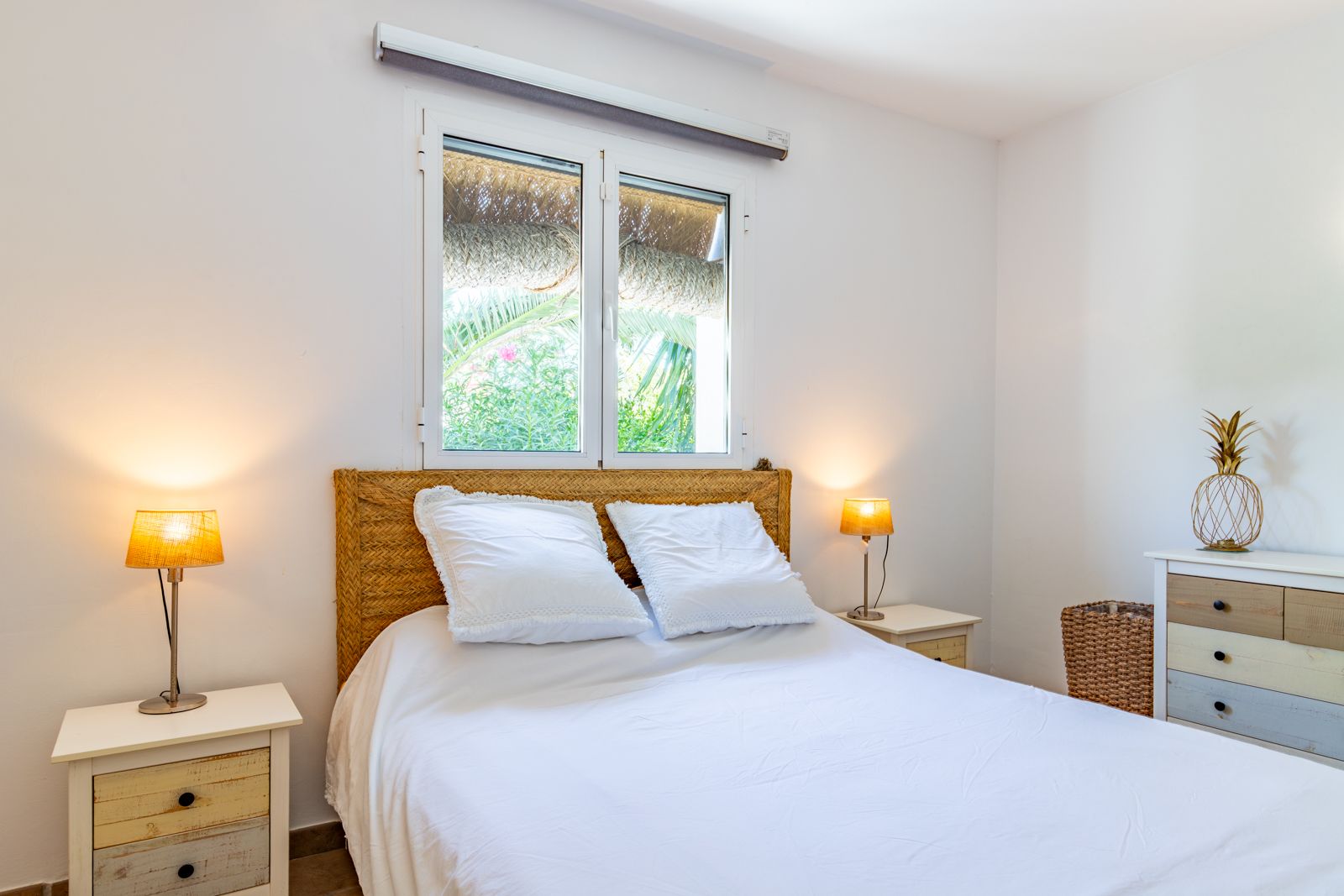Chambre double avec tête de lit en osier, tables de chevet en bois rustique et fenêtre vue jardin. Un espace lumineux au style méditerranéen.