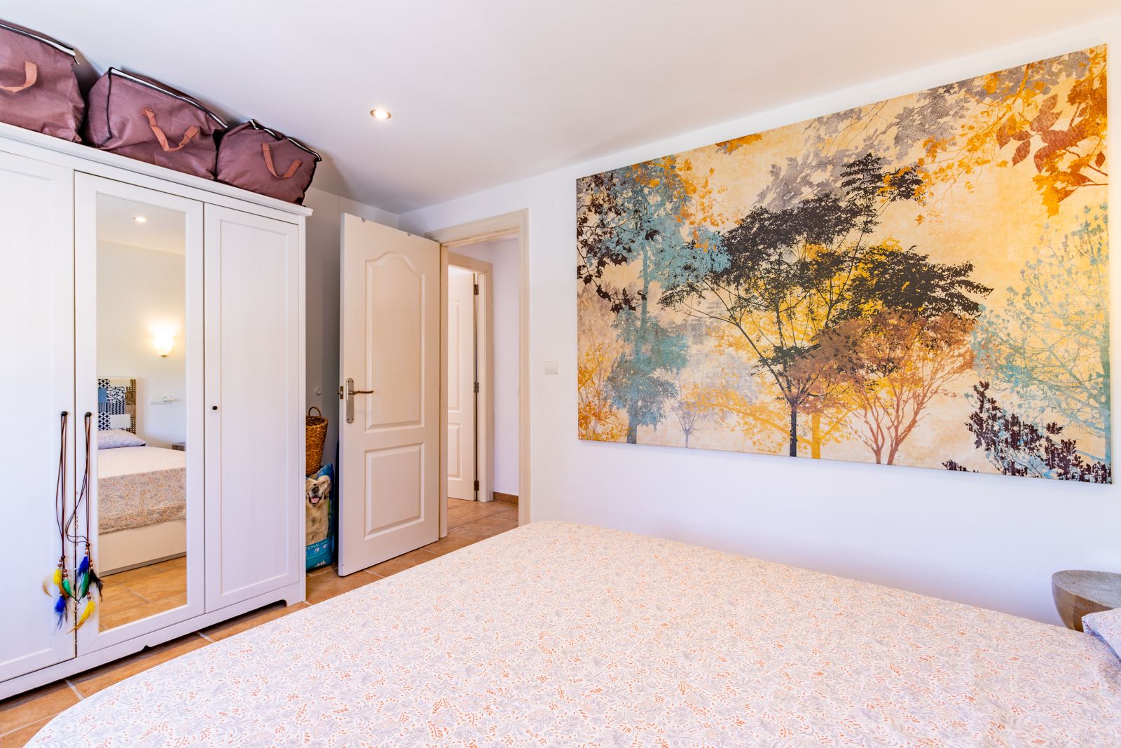 Chambre lumineuse avec armoire blanche à trois portes, miroir central et une grande fresque murale décorative. Carrelage au sol et éclairage encastré.