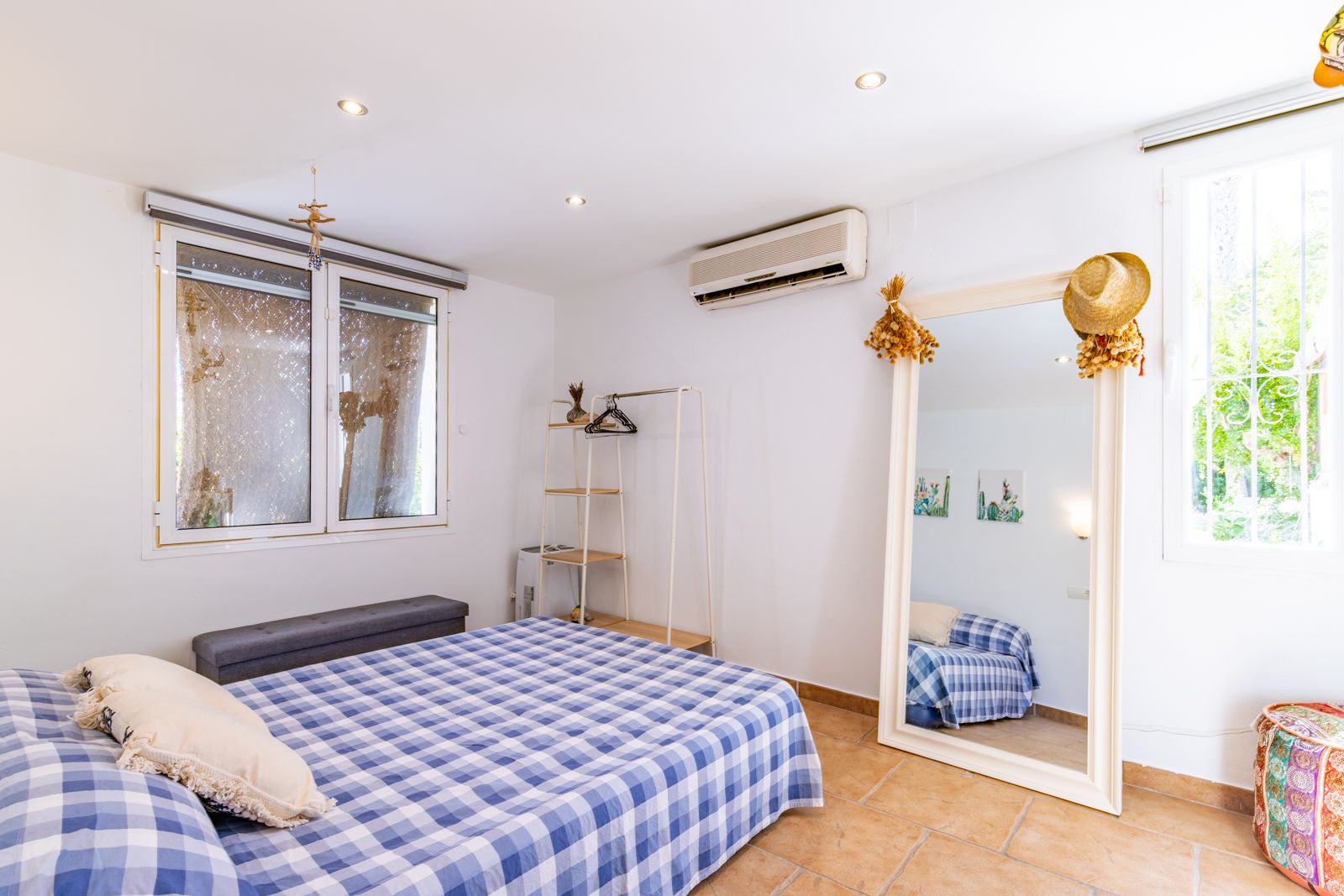 Chambre lumineuse avec sol en terre cuite, climatisation et un grand miroir sur pied. L'espace dispose de spots encastrés et d'une belle luminosité naturelle.