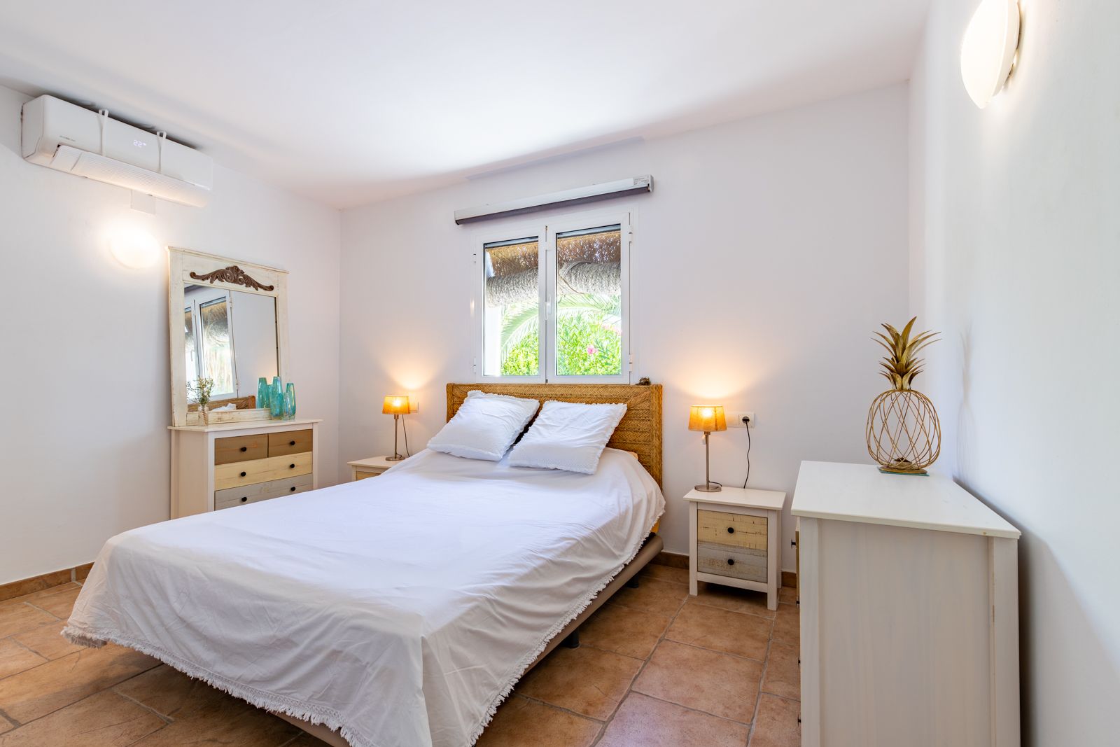 Chambre lumineuse avec sol en terre cuite, climatisation réversible et mobilier rustique. Elle dispose d'une tête de lit en fibres naturelles et d'une fenêtre ouvrant sur la verdure.