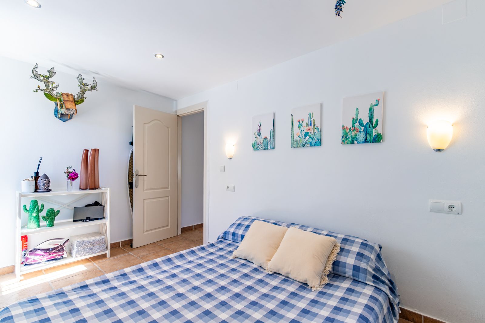 Chambre lumineuse avec sol en terre cuite, lit aux textiles à carreaux bleus et décoration thématique. Éclairage intégré et appliques murales chaleureuses.