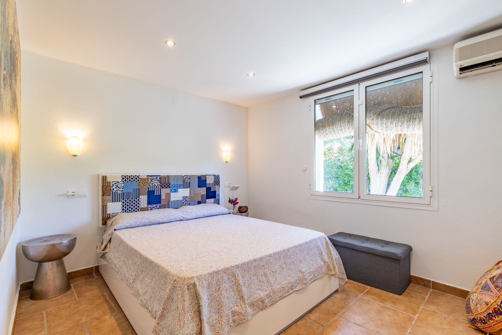 Chambre lumineuse avec sol en terre cuite, tête de lit en patchwork, climatisation et fenêtre offrant une vue sur le jardin.