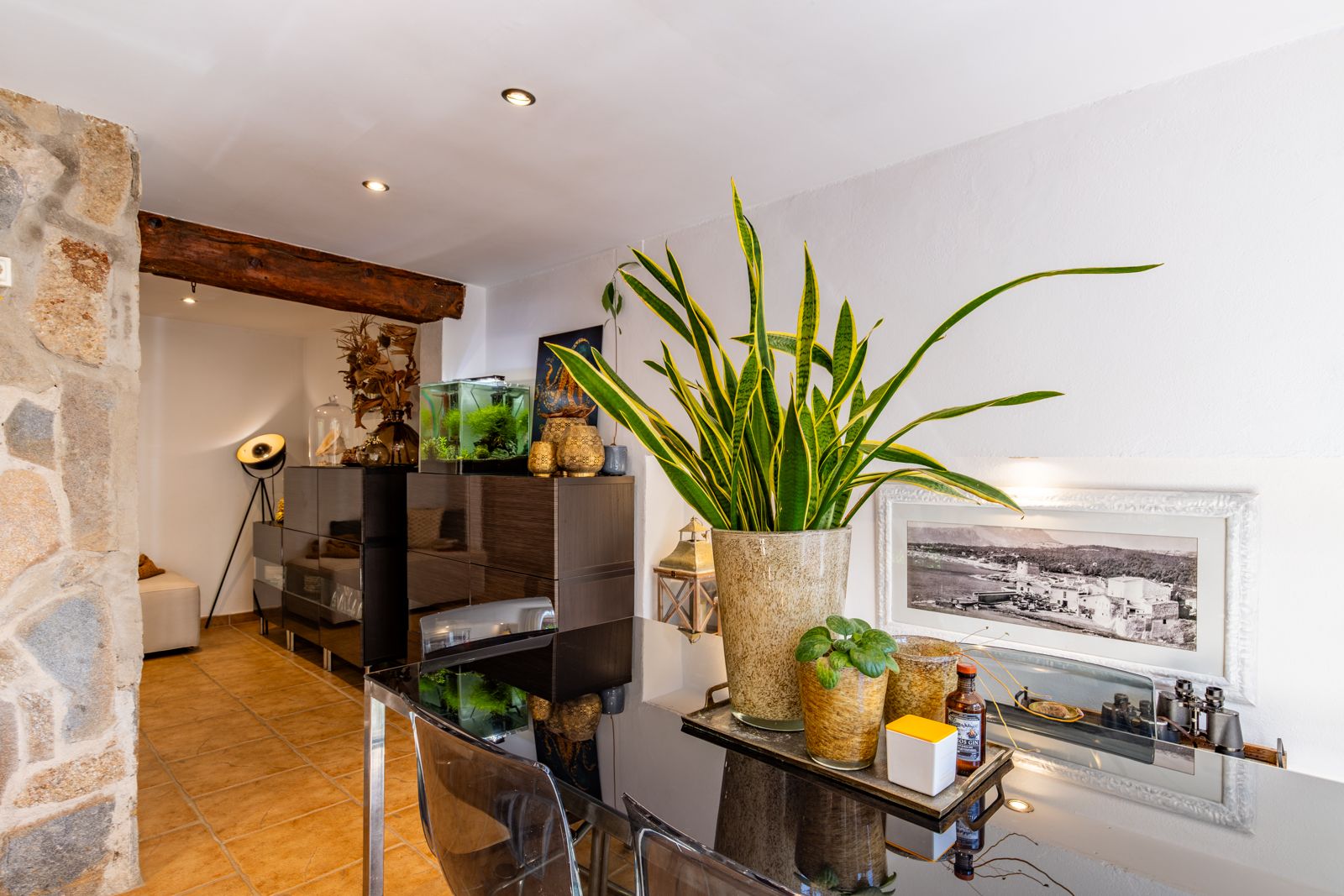 Coin repas avec table en verre noir, chaises transparentes et touches rustiques, incluant une poutre en bois massif et un mur en pierre.