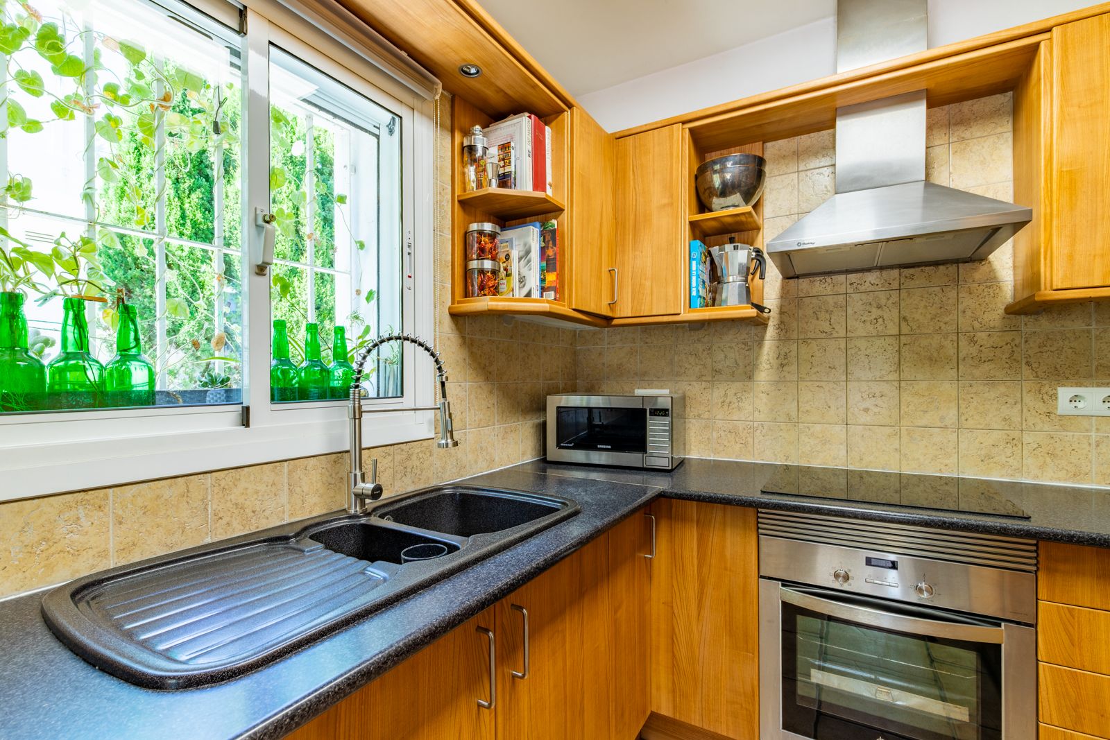 Cuisine équipée avec placards en bois, plan de travail en granit sombre et double évier. Profite de la lumière naturelle et d'appareils en acier inoxydable.